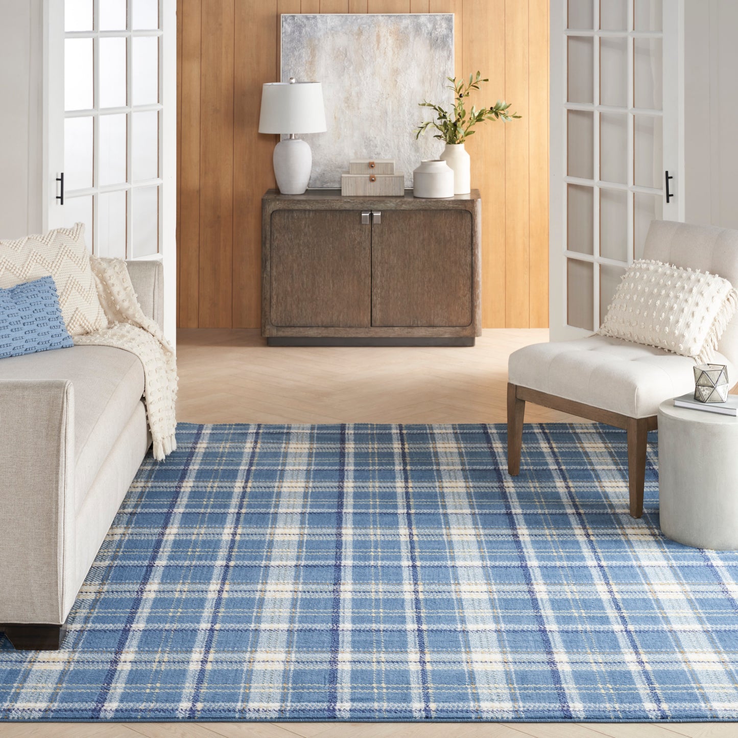 Rustic dark wood sideboard on warm wood wall, blue plaid rug, beige sofa and white armchair in airy living room.