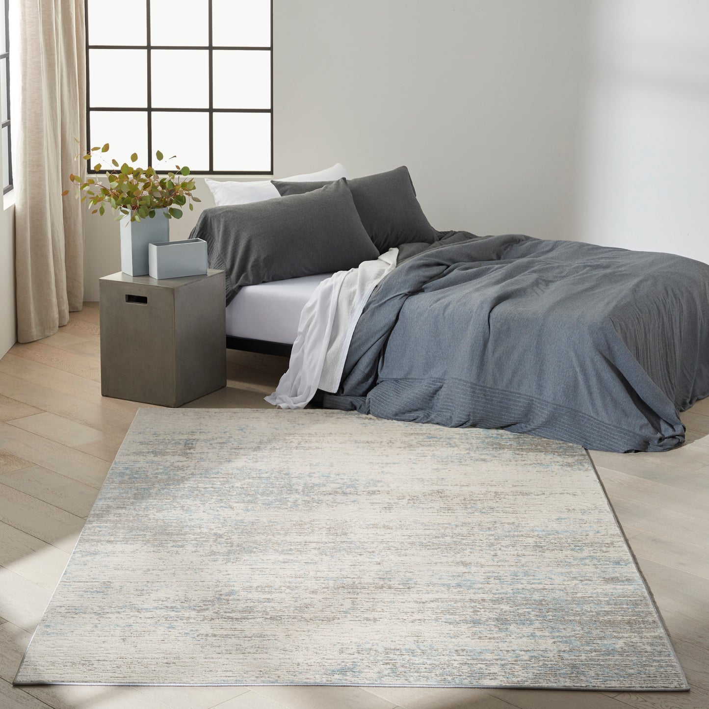 Modern bedroom with gray bedding, white sheets, concrete nightstand, potted plant, and large blue-gray patterned rug.