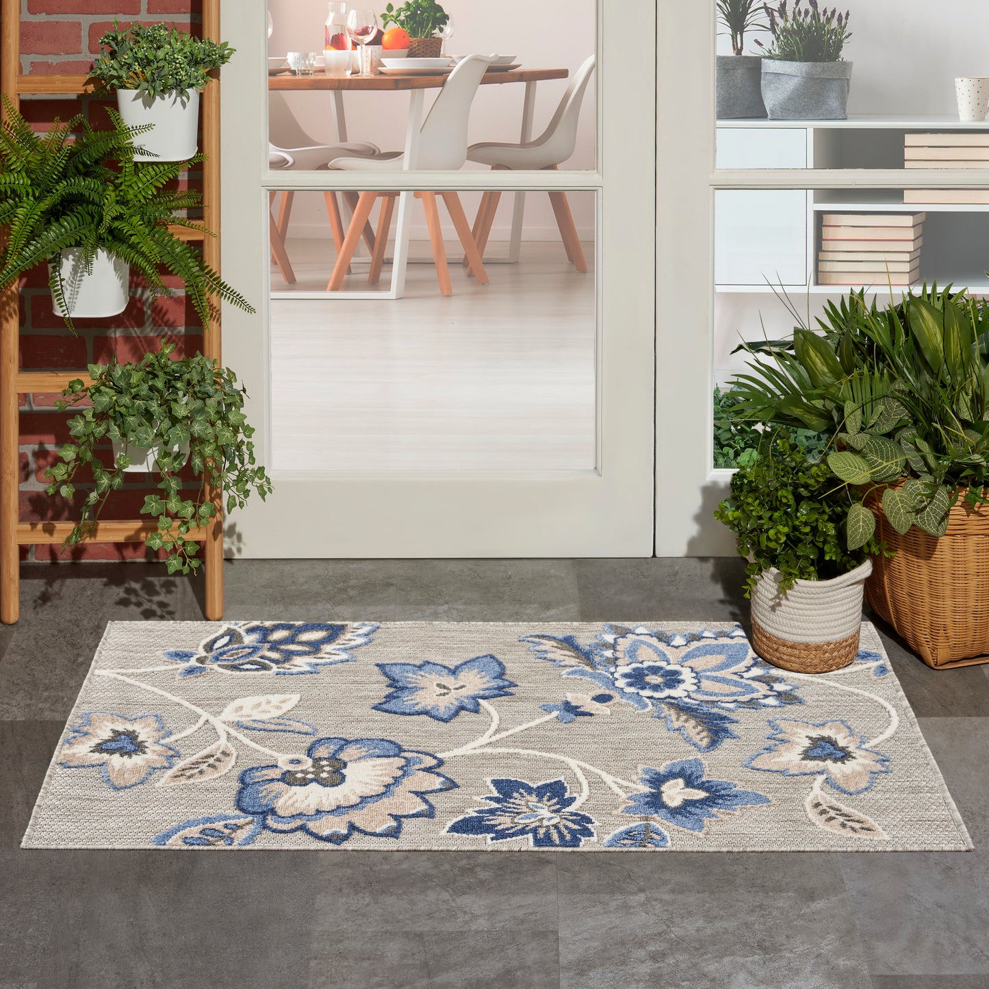 Gray rectangular floral rug with blue flowers at the doorway, flanked by potted plants and a dining room visible inside.