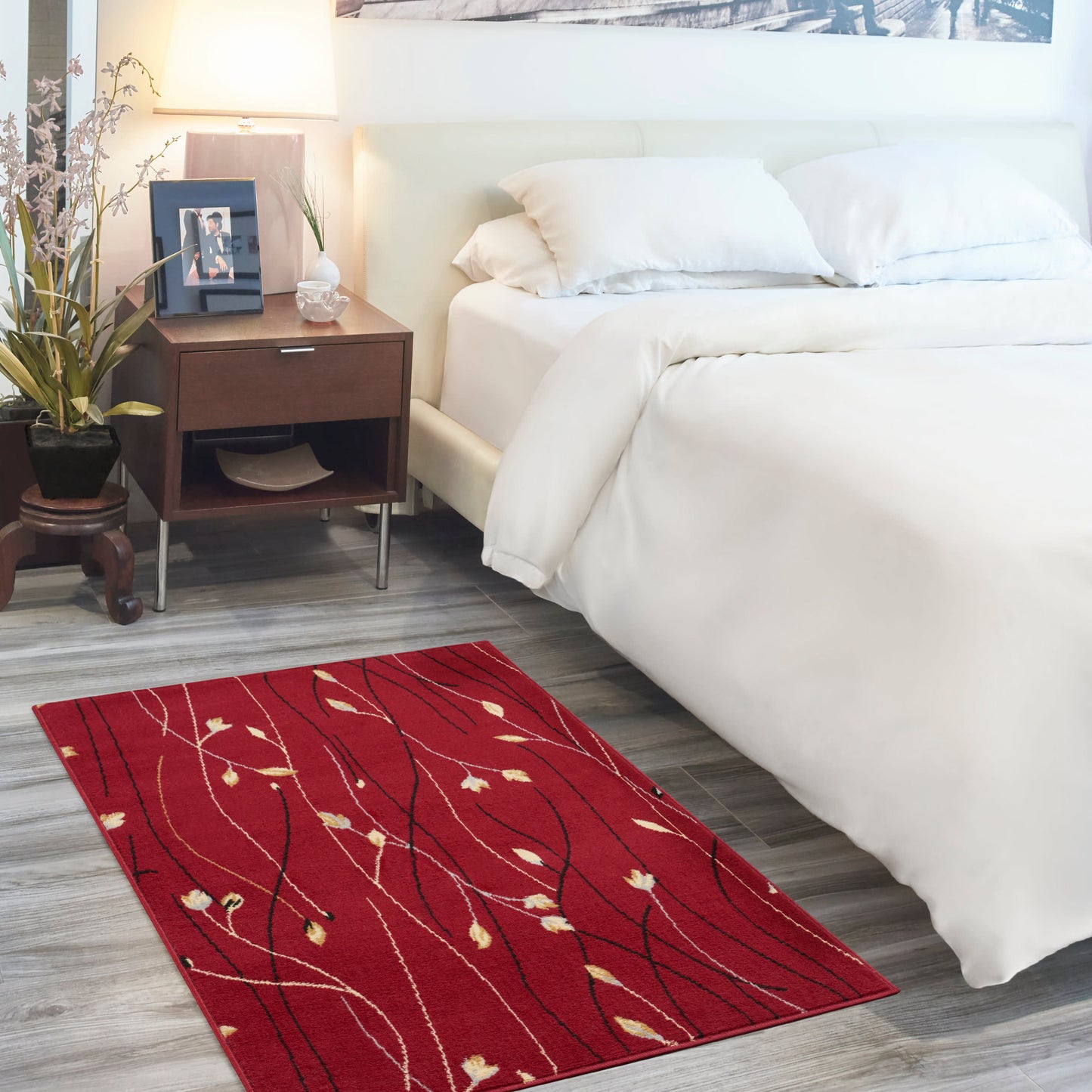 Red rectangular area rug with white leaf vines on gray wood floor beside a white bed; dark wood nightstand & lamp.