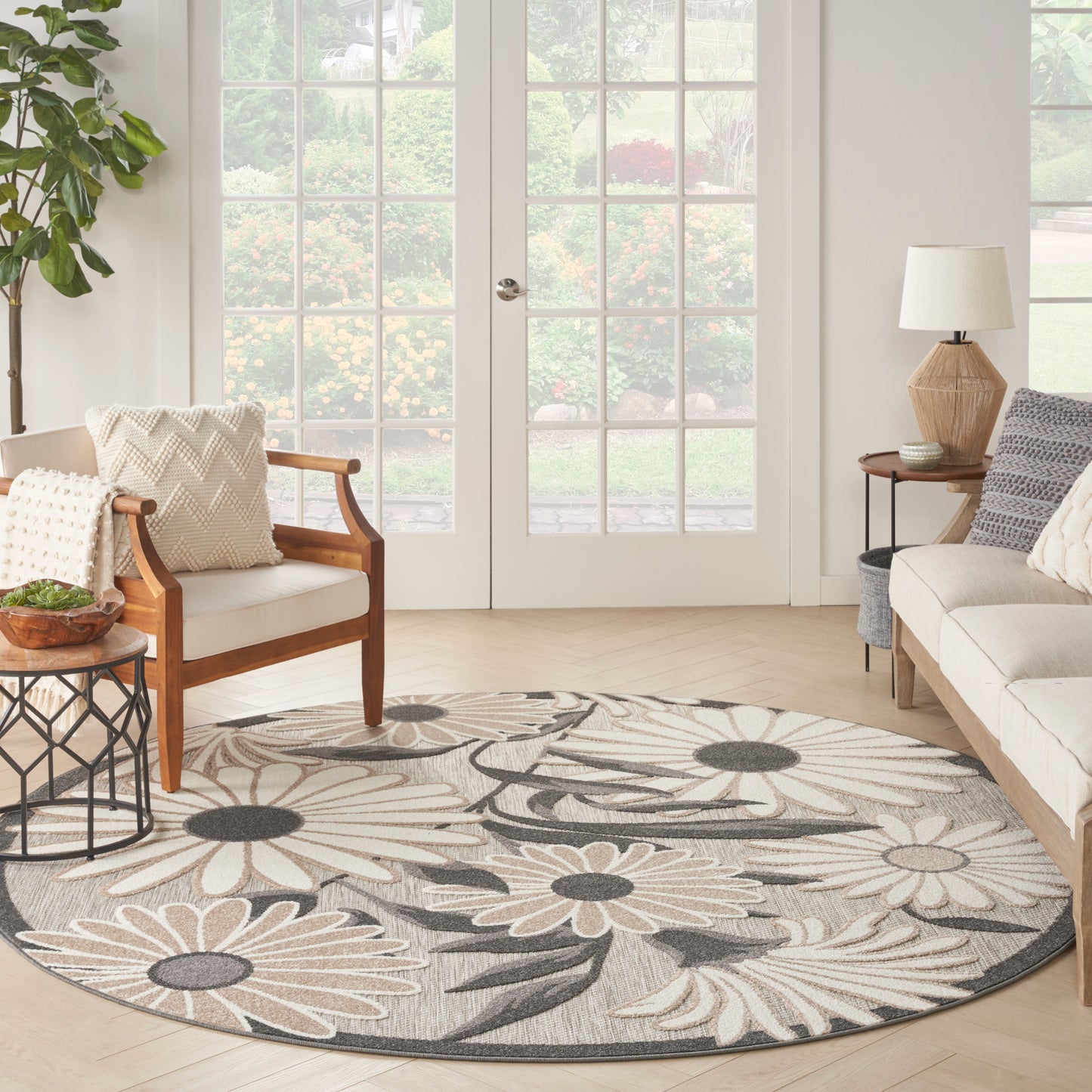 Round floral rug in beige and gray under a light living room with wood armchairs, neutral sofa, plant, and French doors.