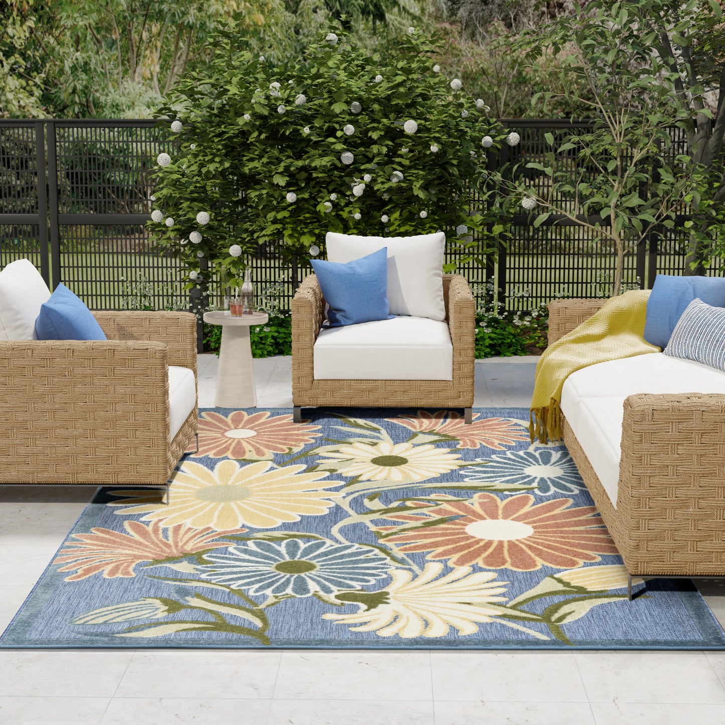 Outdoor seating area with wicker furniture, white cushions, blue accents, and a bold floral rug in blue, yellow, orange.