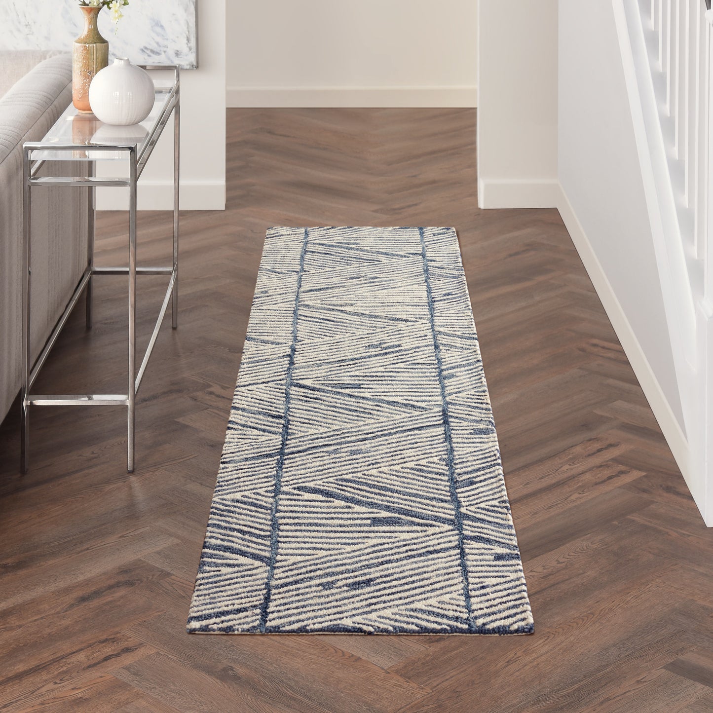 Blue geometric hallway runner rug on dark herringbone floor beside a glass console table with vases.