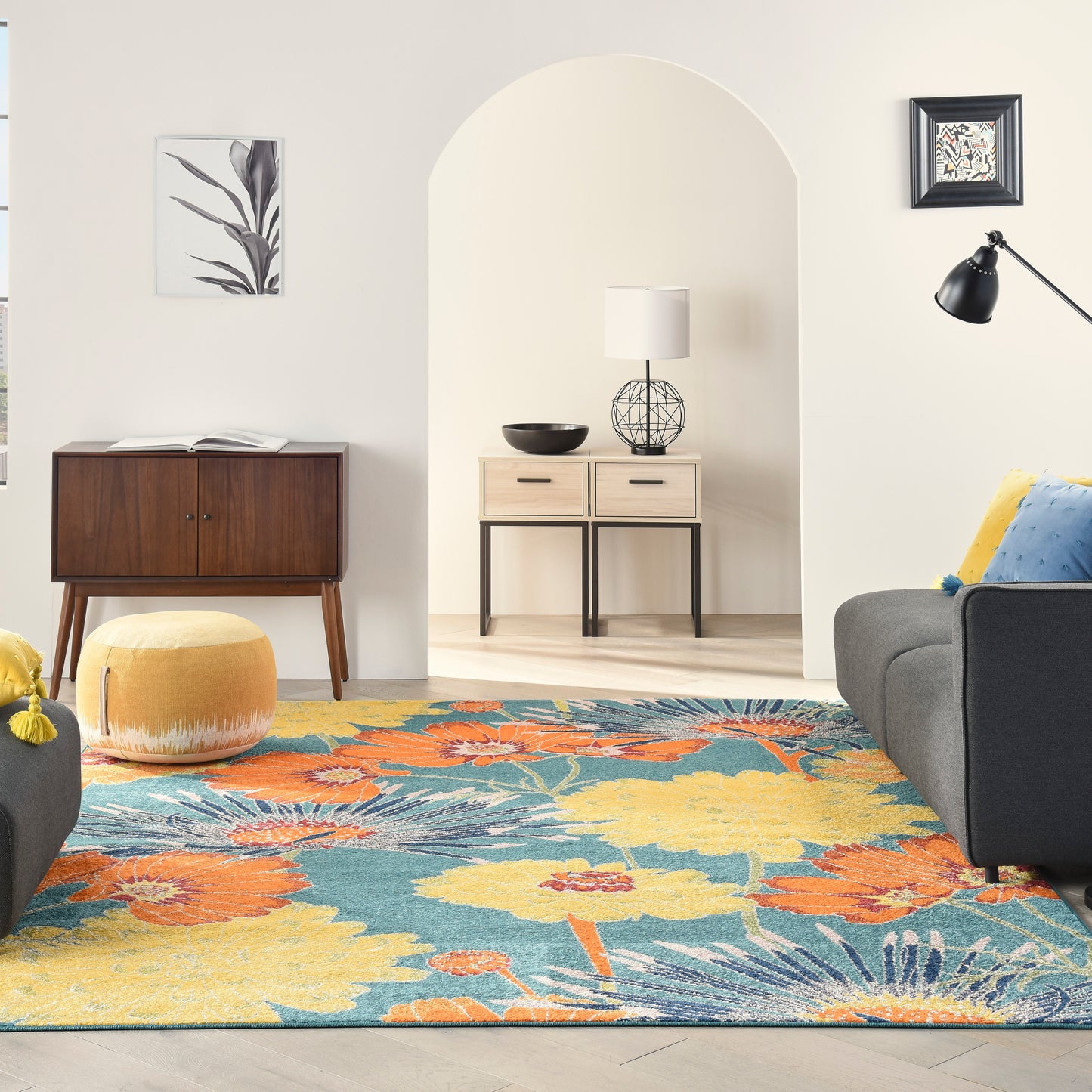 Living room with vibrant floral rug, mid-century furniture, yellow pouf, grey sofa, arched doorway, wood cabinet, floor lamp.