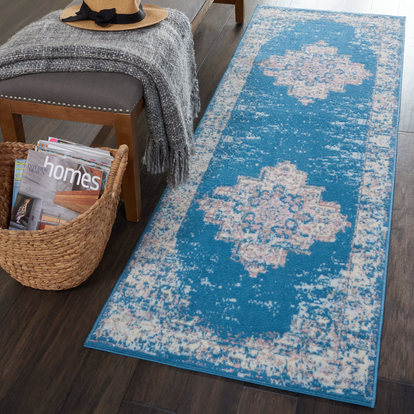Blue distressed runner rug with pale pink medallions on dark wood floor beside bench with a gray throw and wicker magazines.