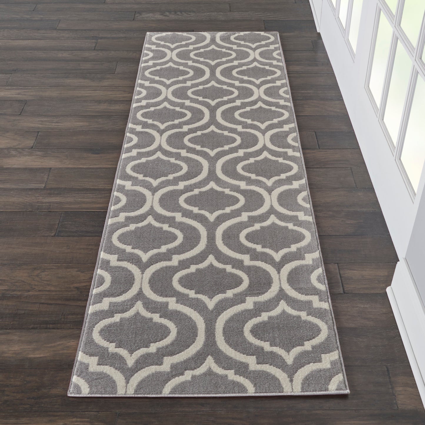 Grey & beige trellis-patterned 7' long runner rug on dark wood flooring beside a glass-paneled wall.