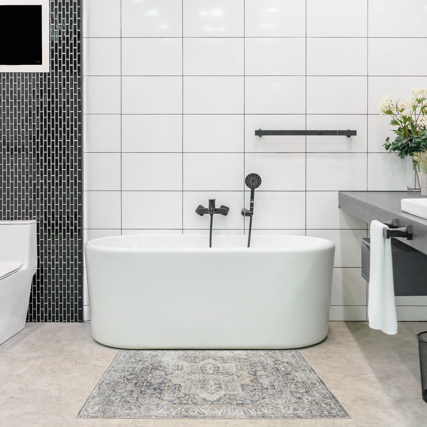 White freestanding oval bathtub with black hardware & handheld shower, white tile walls, gray rug, modern bathroom.