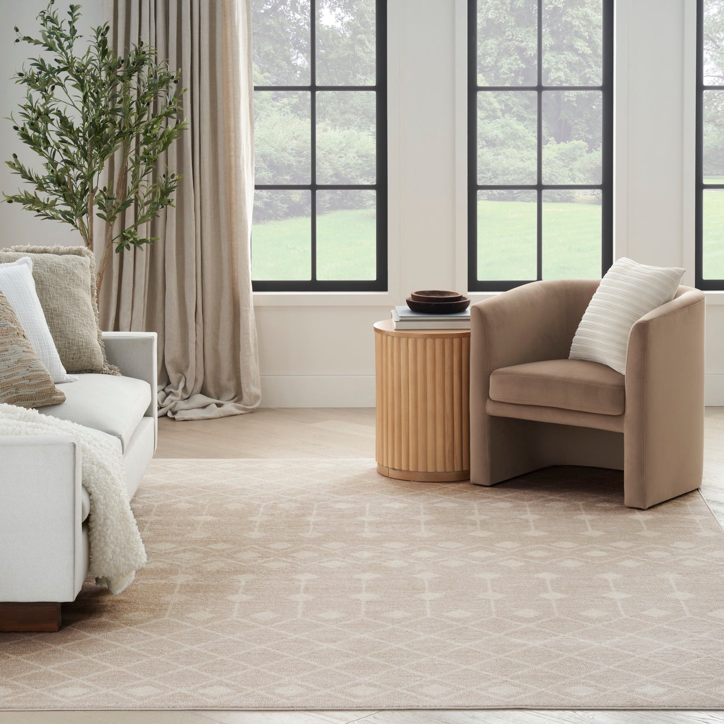 Neutral living room with beige armchair, white sofa, round wood side table, light patterned rug, plant, and grid windows.