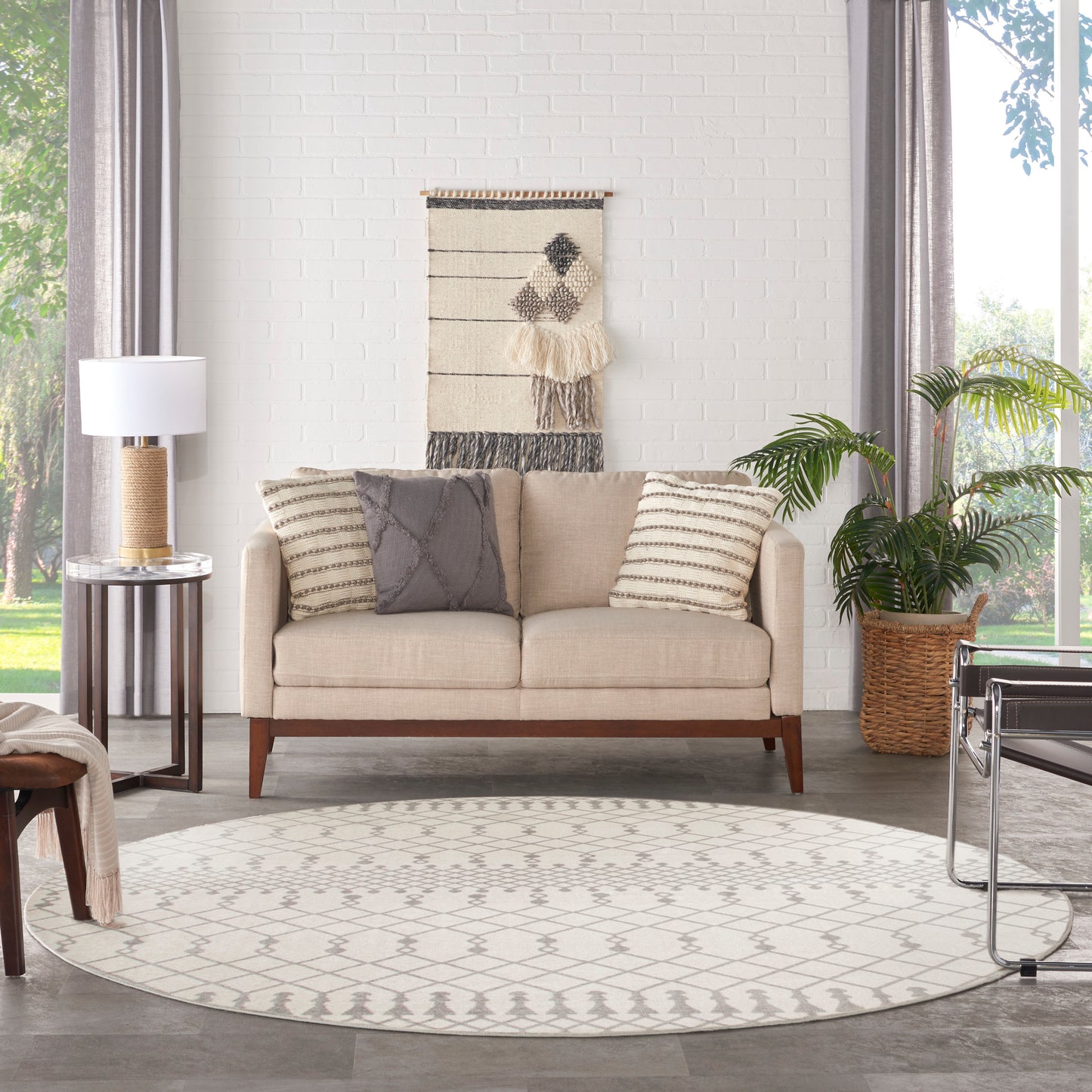 Beige mid-century sofa with striped and grey cushions on a round geometric rug; woven wall hanging, lamp, and potted plant.