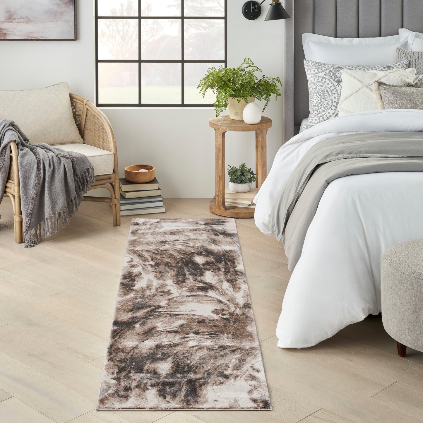 Long brown-beige abstract runner rug on light wood floor beside a white bed, wicker chair, and potted plant.