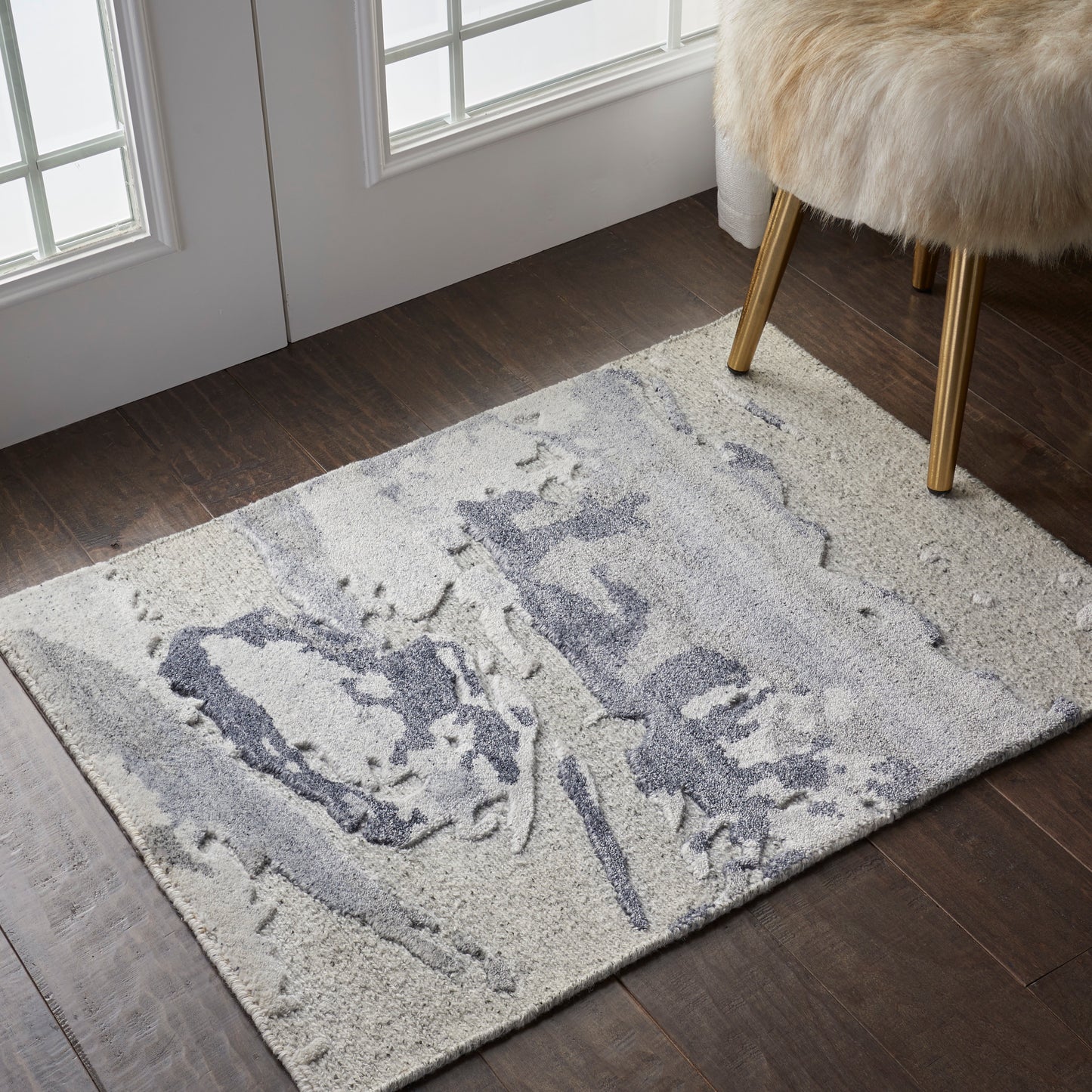 Cream shag area rug with abstract gray-blue shapes on a dark wood floor, beside a fur-covered stool with gold legs.