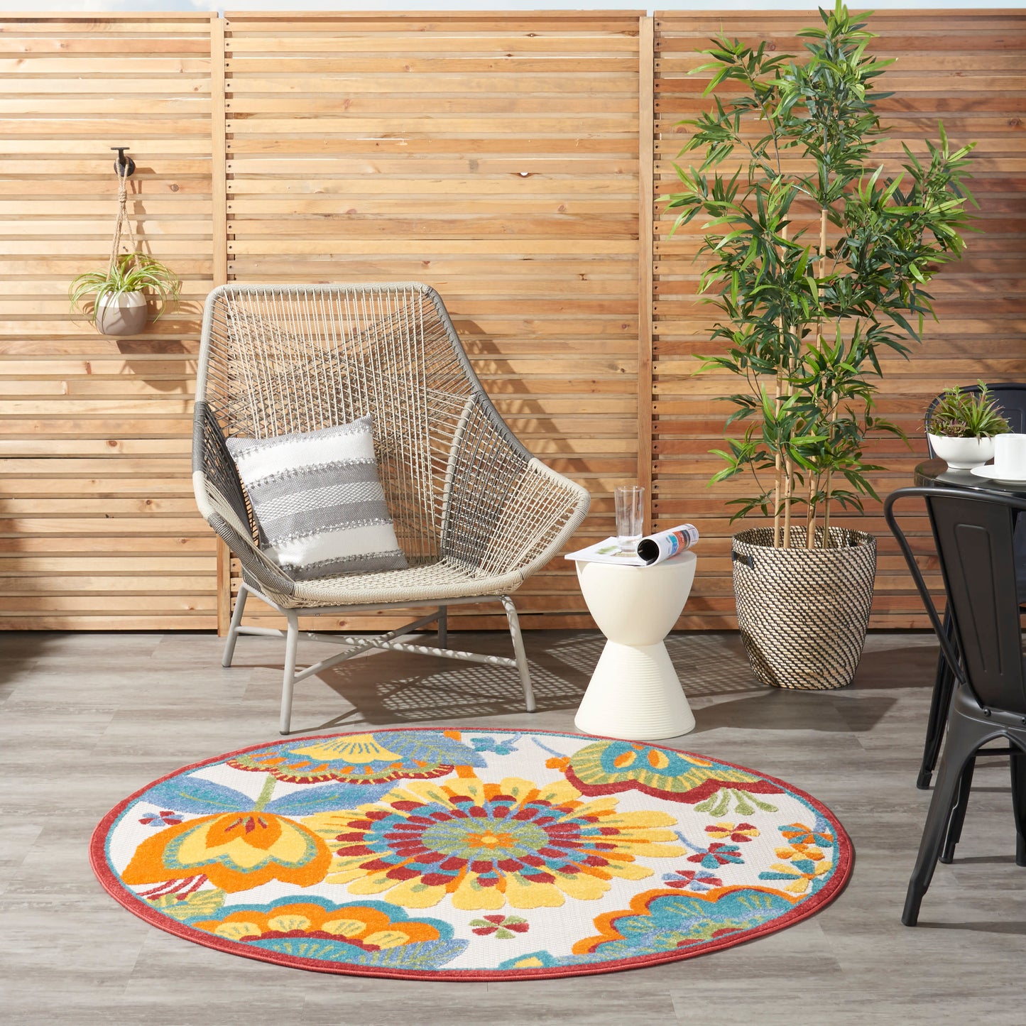 Outdoor patio: wooden slat fence, woven rattan chair with striped cushion, round colorful floral rug, potted plants.