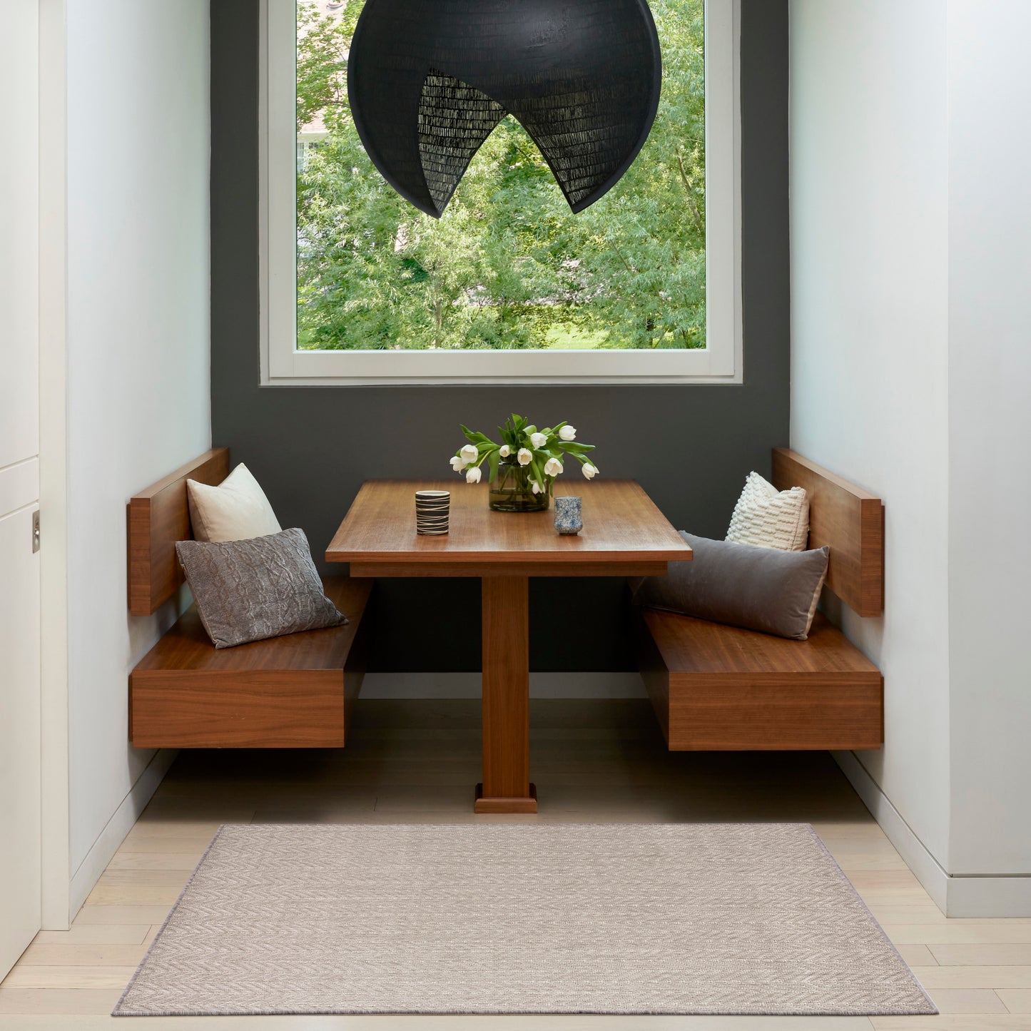 Wooden corner dining nook with pedestal table, built-in benches, gray walls, black pendant, beige rug, cushions, flowers.