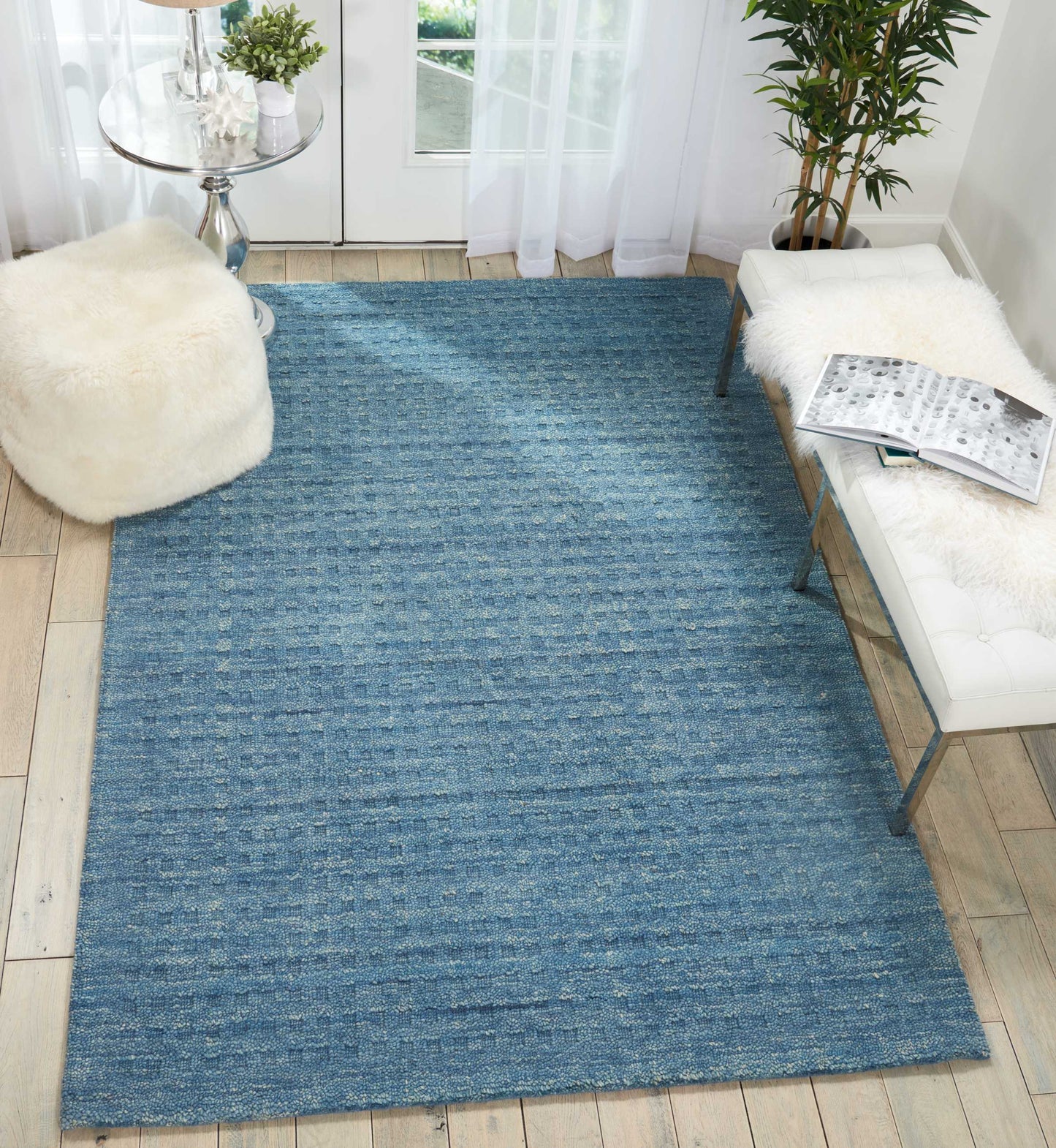 Blue textured woven area rug on light wood floor with white tufted bench, sheepskin throw, round glass table, and potted plants.