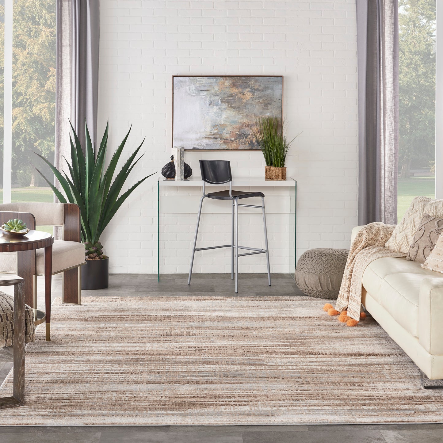 Modern living room with glass console, black metal bar stool, white brick wall, beige sofa, large plant, neutral striped rug.