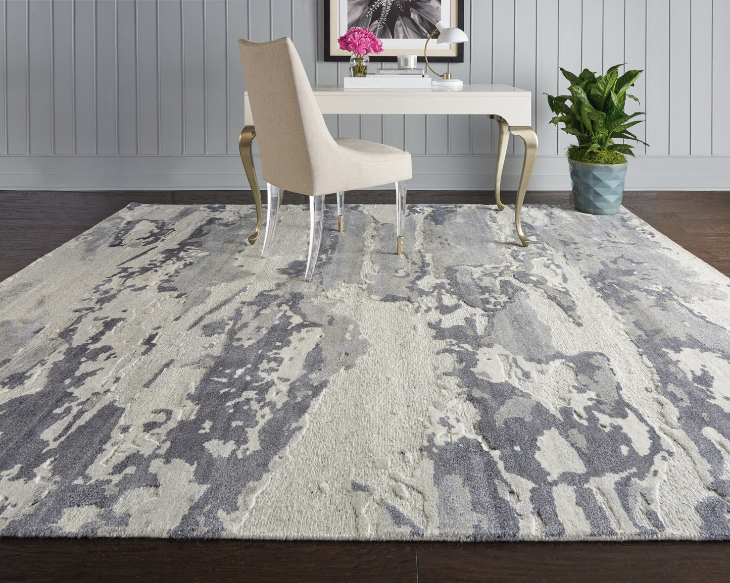 Beige upholstered desk chair with clear acrylic legs, white desk with gold curved legs, blue-gray abstract rug, potted plant.