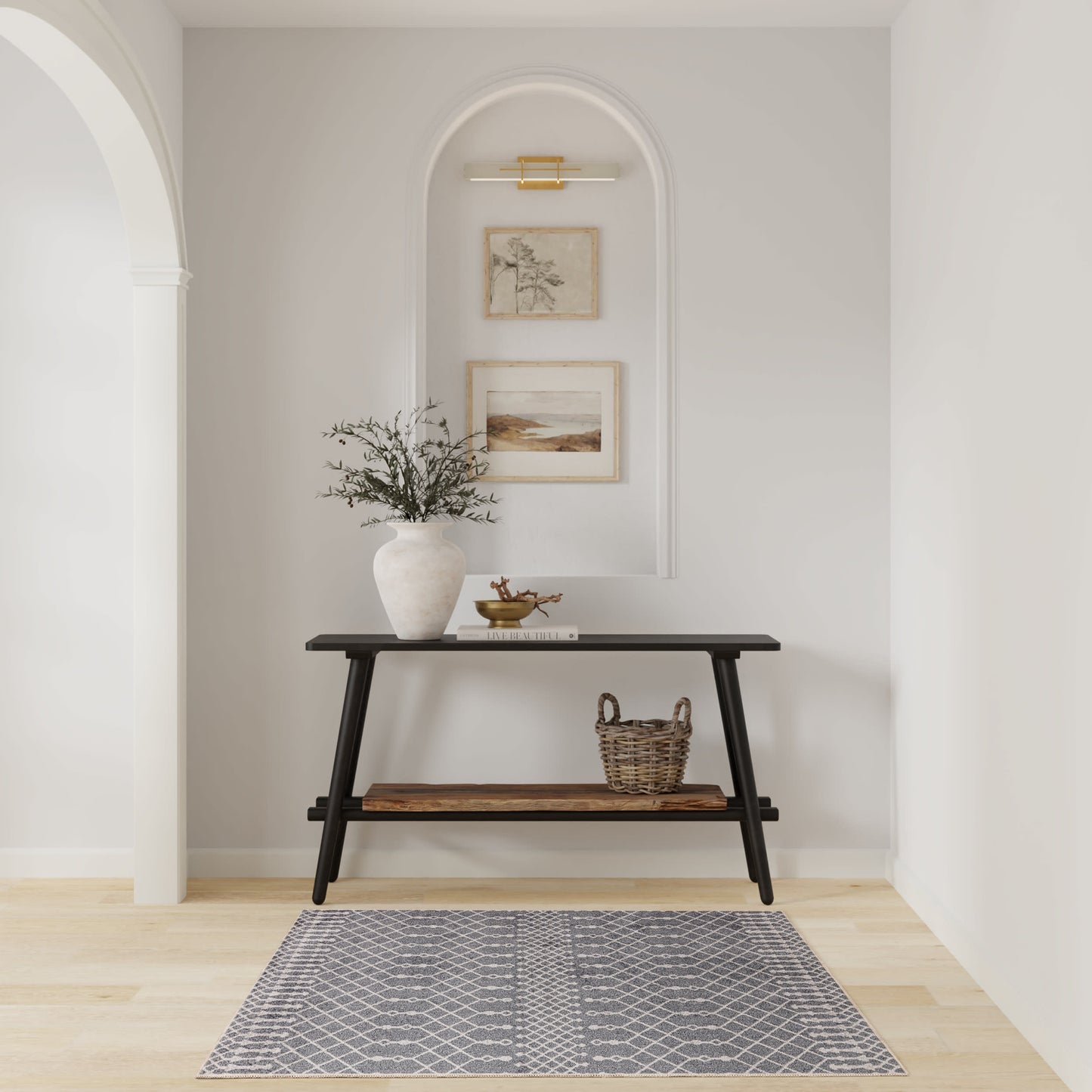 Black wooden two-tier console table in a pale entry alcove with a vase of greenery, woven basket, arch niche with framed art, and a grey patterned rug.