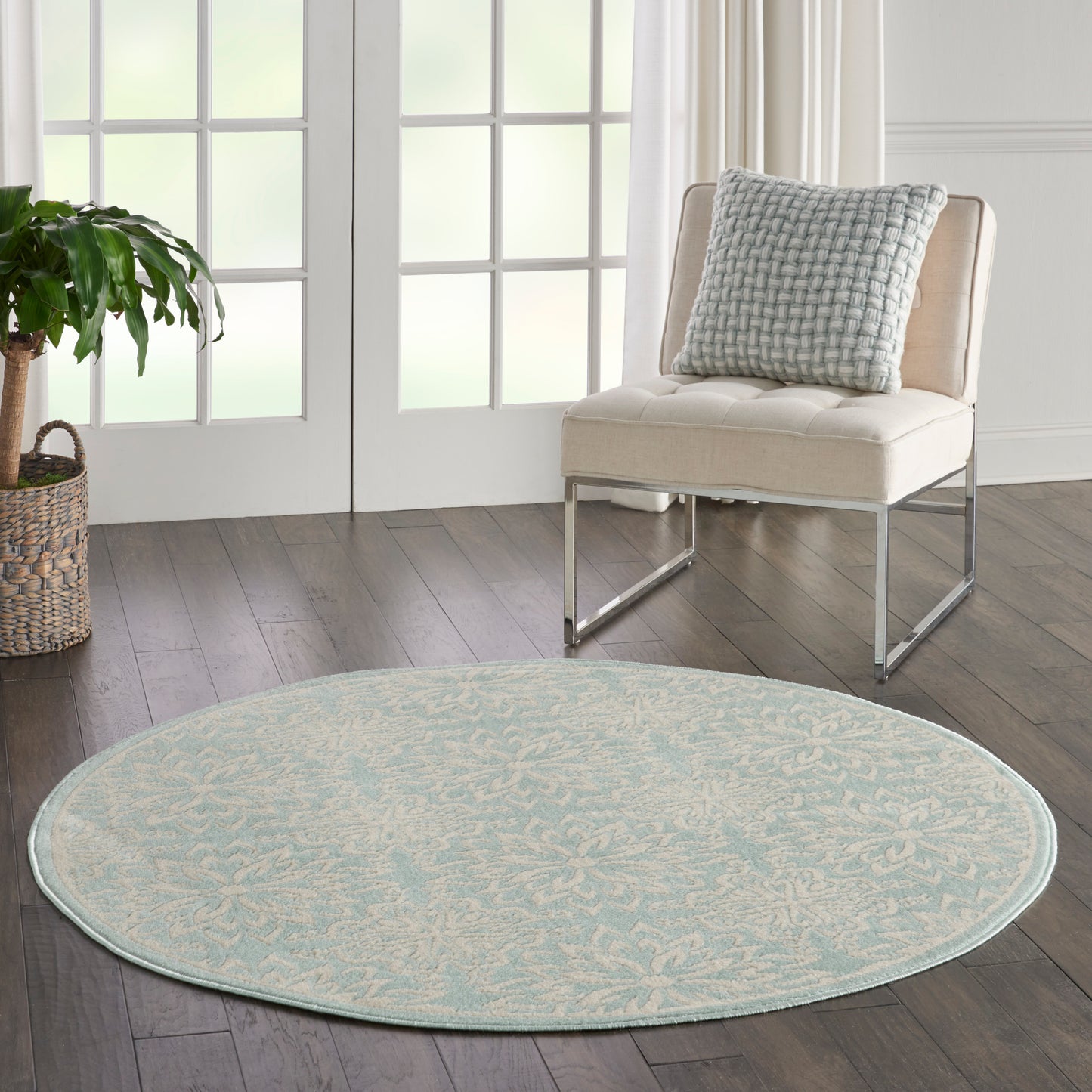 Round light blue rug with subtle beige floral motif on dark wood floor; modern beige chair with blue patterned cushion.
