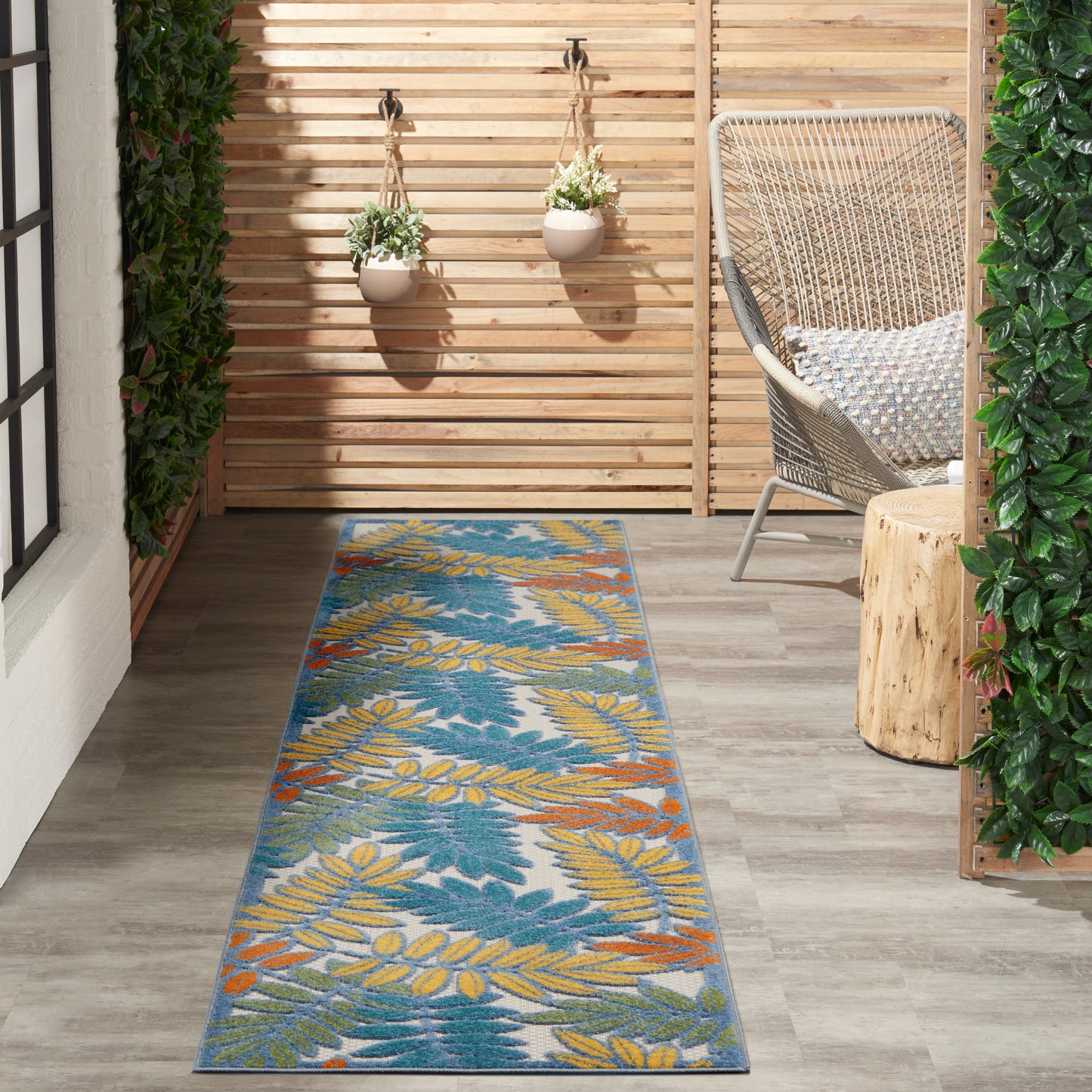 Outdoor rug runner with blue background and yellow-leaf print beside a wicker chair, wooden slat fence and hanging planters.