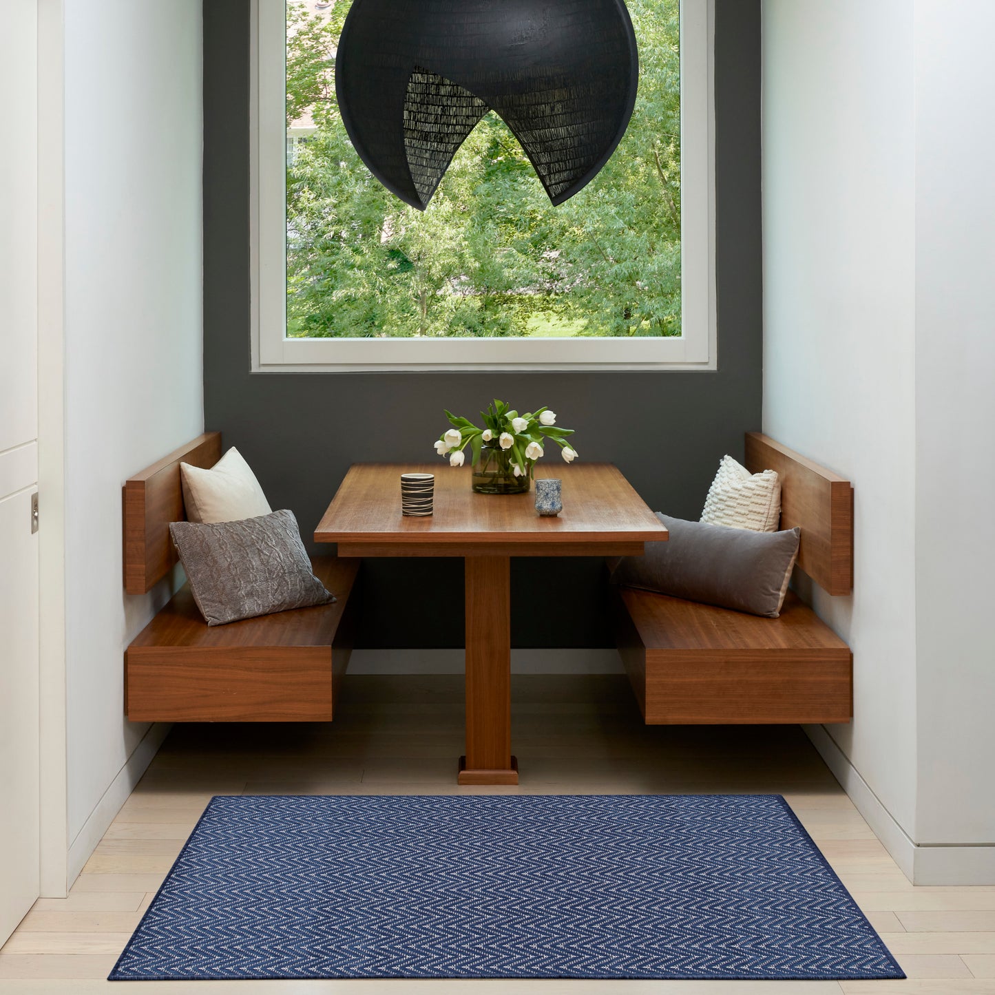 Built-in wooden dining nook with two benches, square table, navy chevron rug, black pendant lamp, and flowers by window.