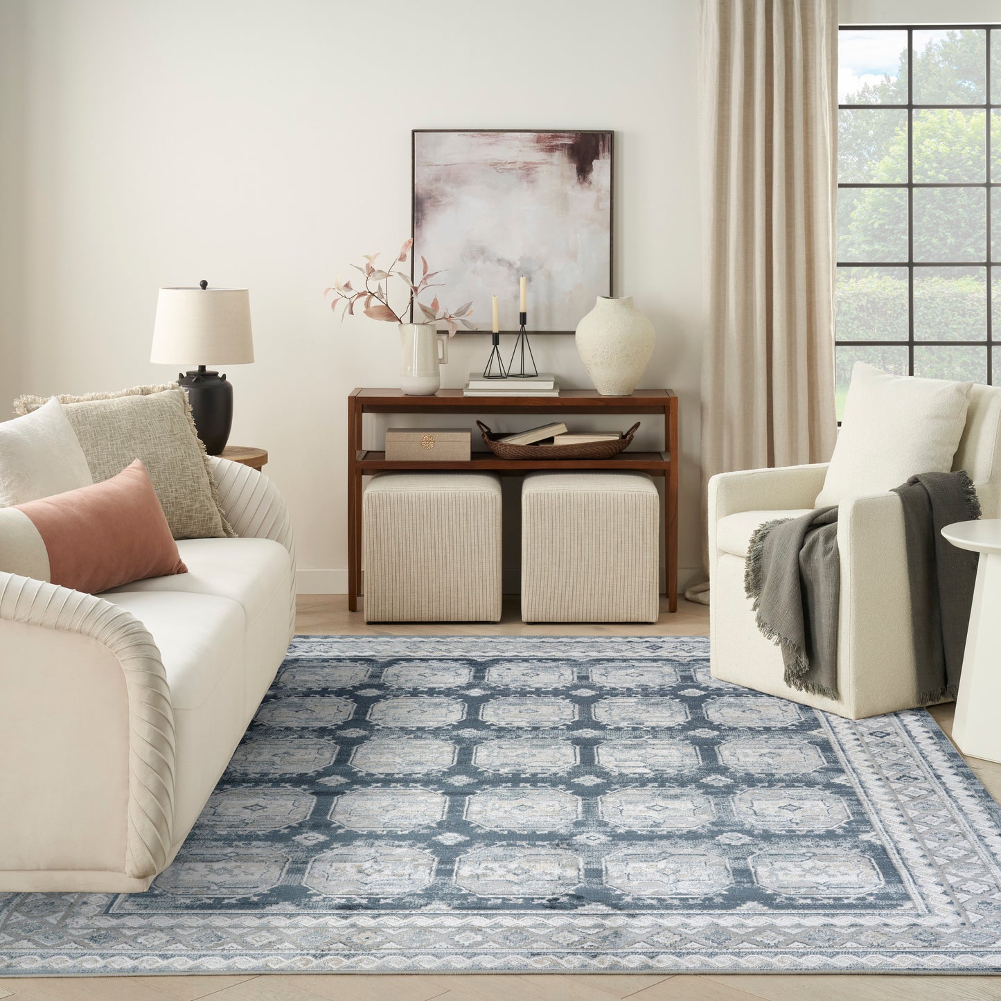 Cozy living room with cream sofa and armchair, blue patterned rug, wooden console with vases and art, beige ottomans.