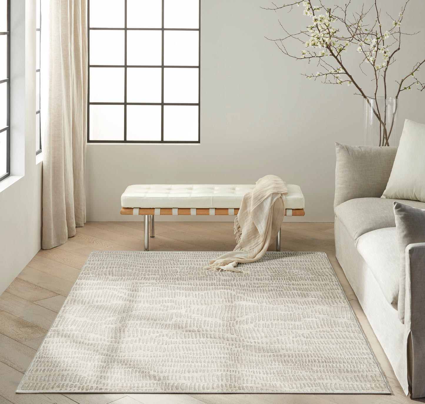 Minimalist living room with a tufted white bench on metal legs, beige throw, ivory rug, light wood floors, and gray sofa.