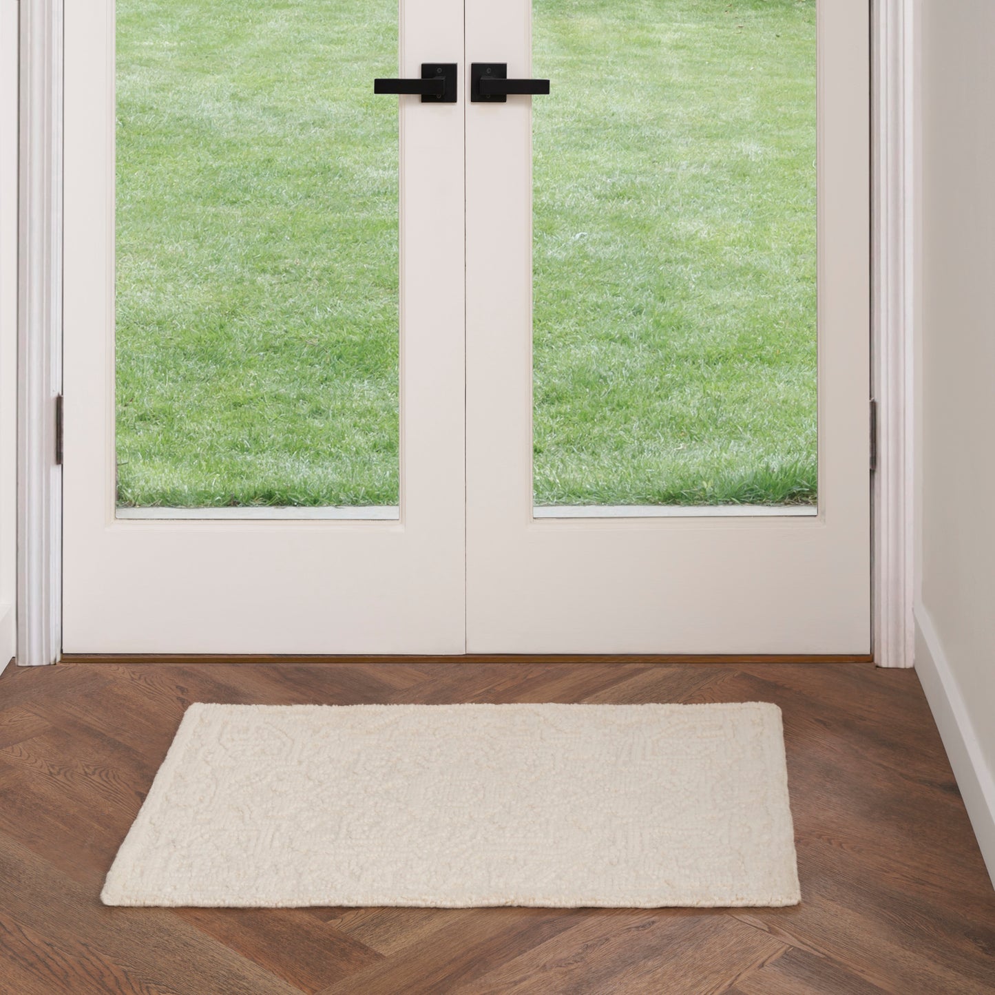 Cream plush entry mat on dark wood herringbone floor in front of white double doors with glass panes and black handles.