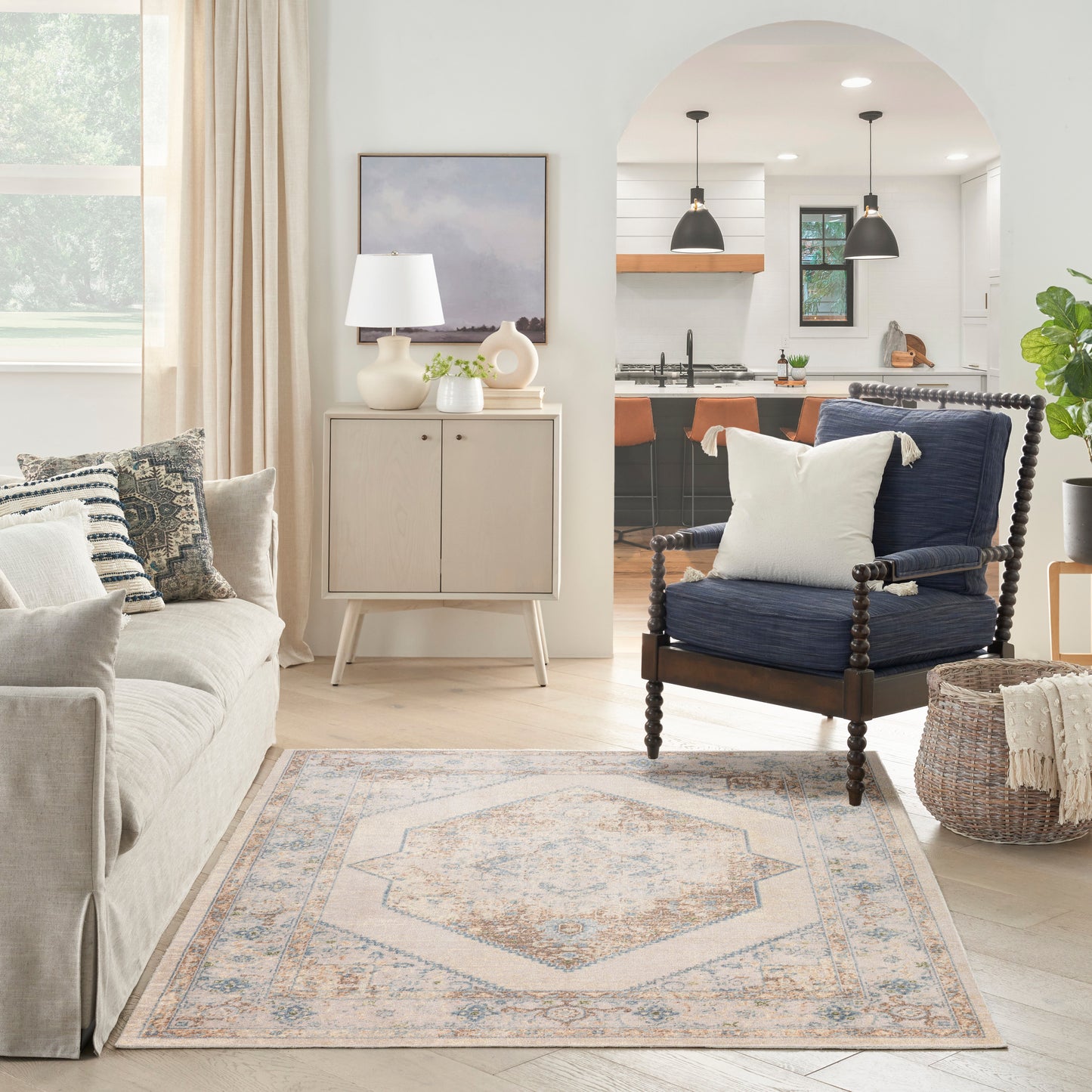 Bright living room with a beige fabric sofa, cream cabinet, navy wood-frame armchair, pastel medallion rug, woven basket, and open kitchen arch.