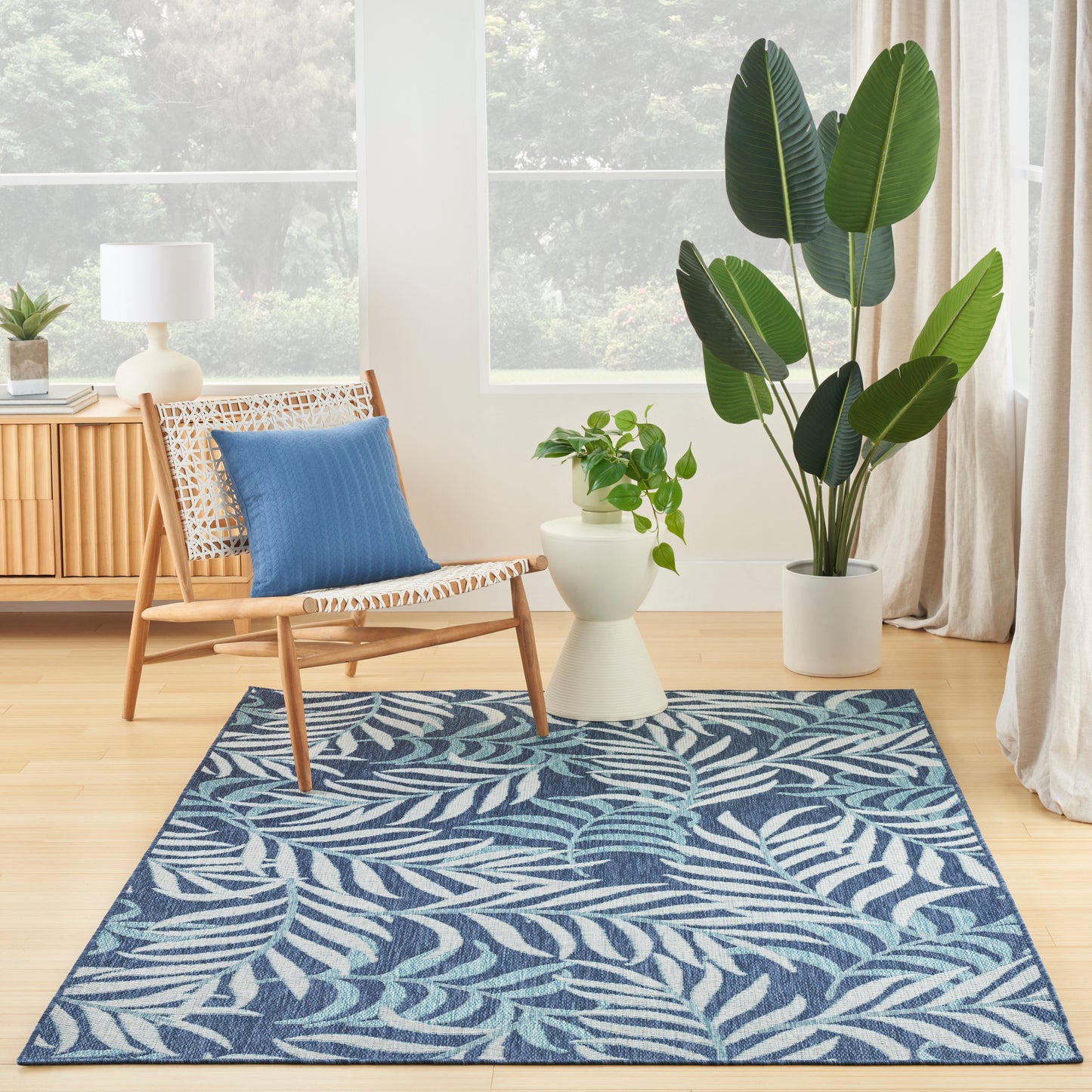 Blue leaf-pattern rug on light wood floor with wicker chair, blue cushion, white side table, and tall potted plants.