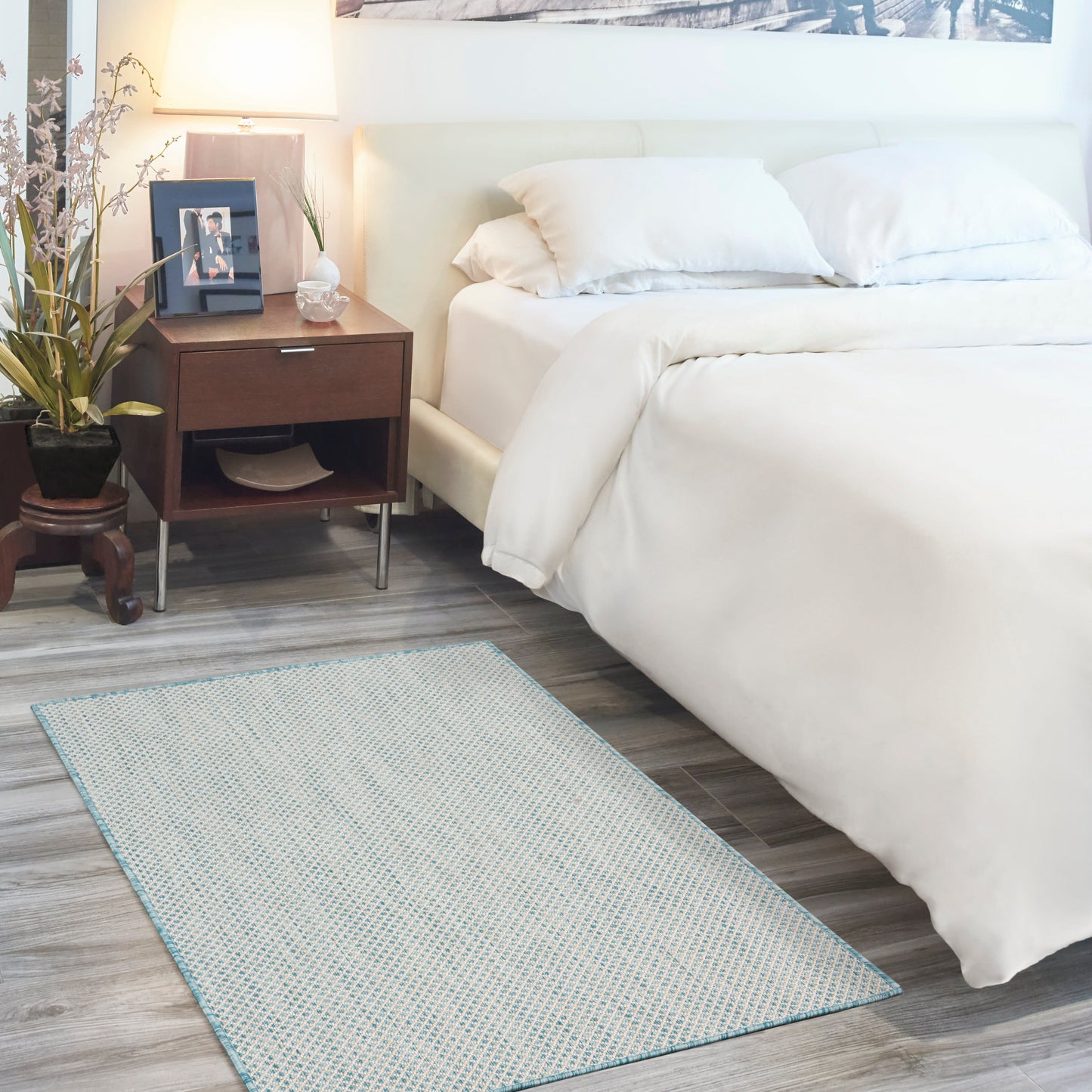 Light blue patterned rug on the floor in a bedroom scene with a white bed, thick comforter and two pillows, a dark wood nightstand with a lamp, plant and photo frame.