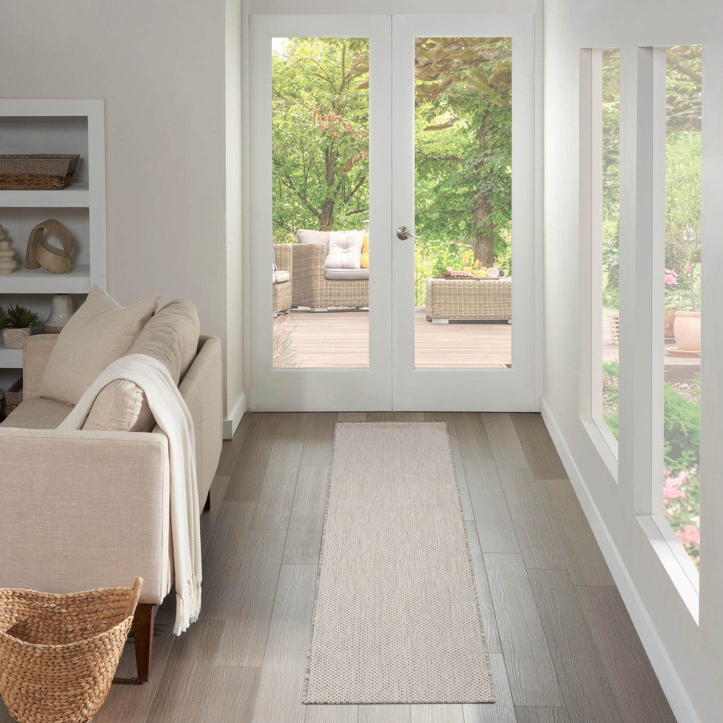 Neutral living room with beige sofa, light wood floors, white shelving, and a taupe runner rug along the hallway to doors.