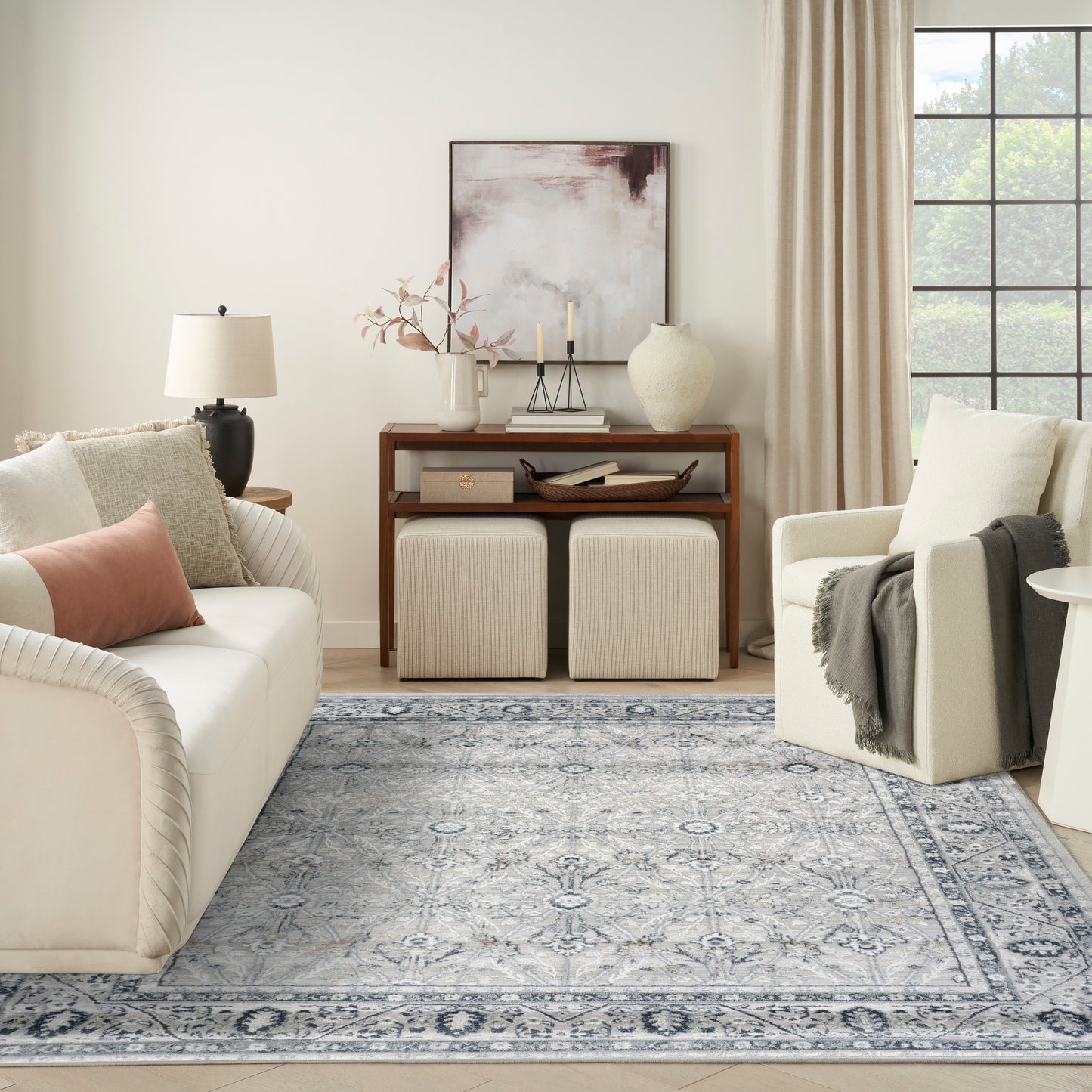 Neutral living room with cream sofa, beige armchair, blue rug, wooden console and ottomans, vase, lamp, abstract art.