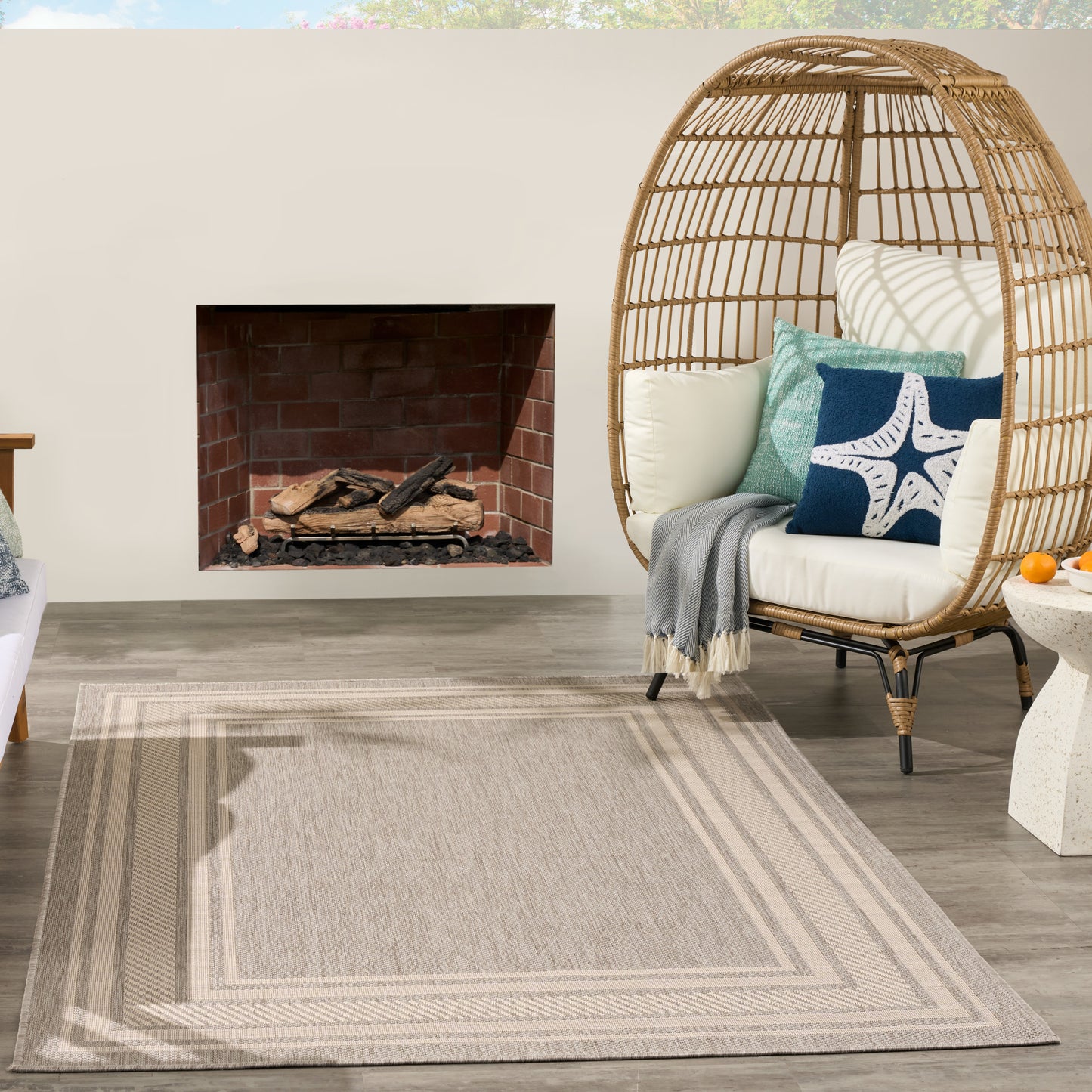 Outdoor patio with brick fireplace, round woven wicker egg chair, cream cushions, blue pillows, and a beige striped rug.