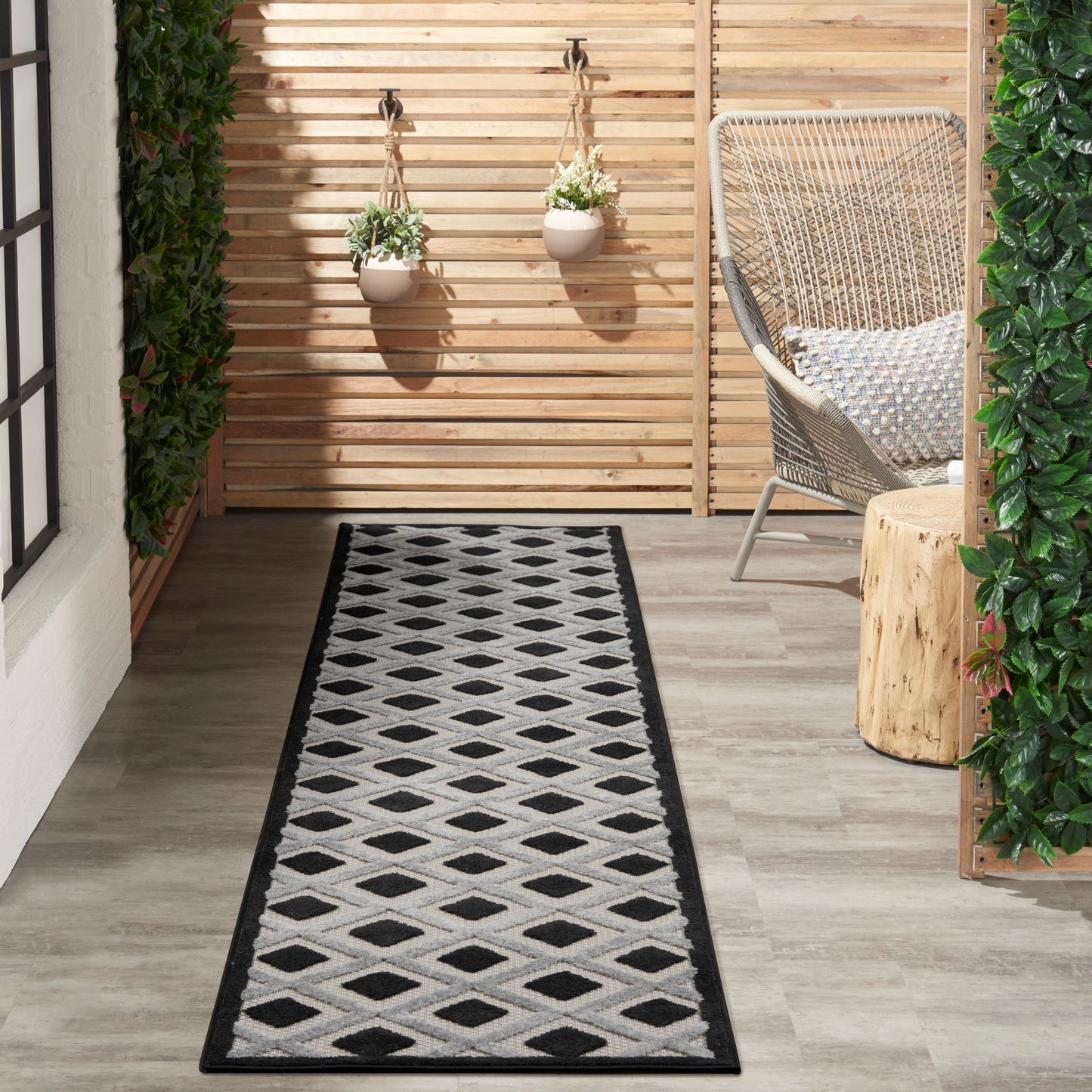 Runner rug with a black diamond pattern on a gray wooden floor beside a wicker chair and potted plants.