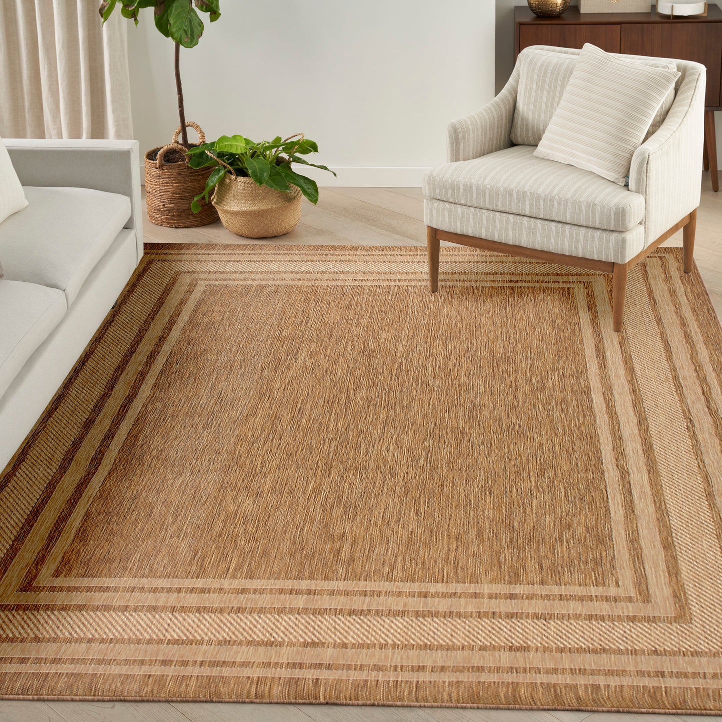 Natural jute area rug with double border in tan and brown; cream striped armchair and leafy plants in a cozy living room.