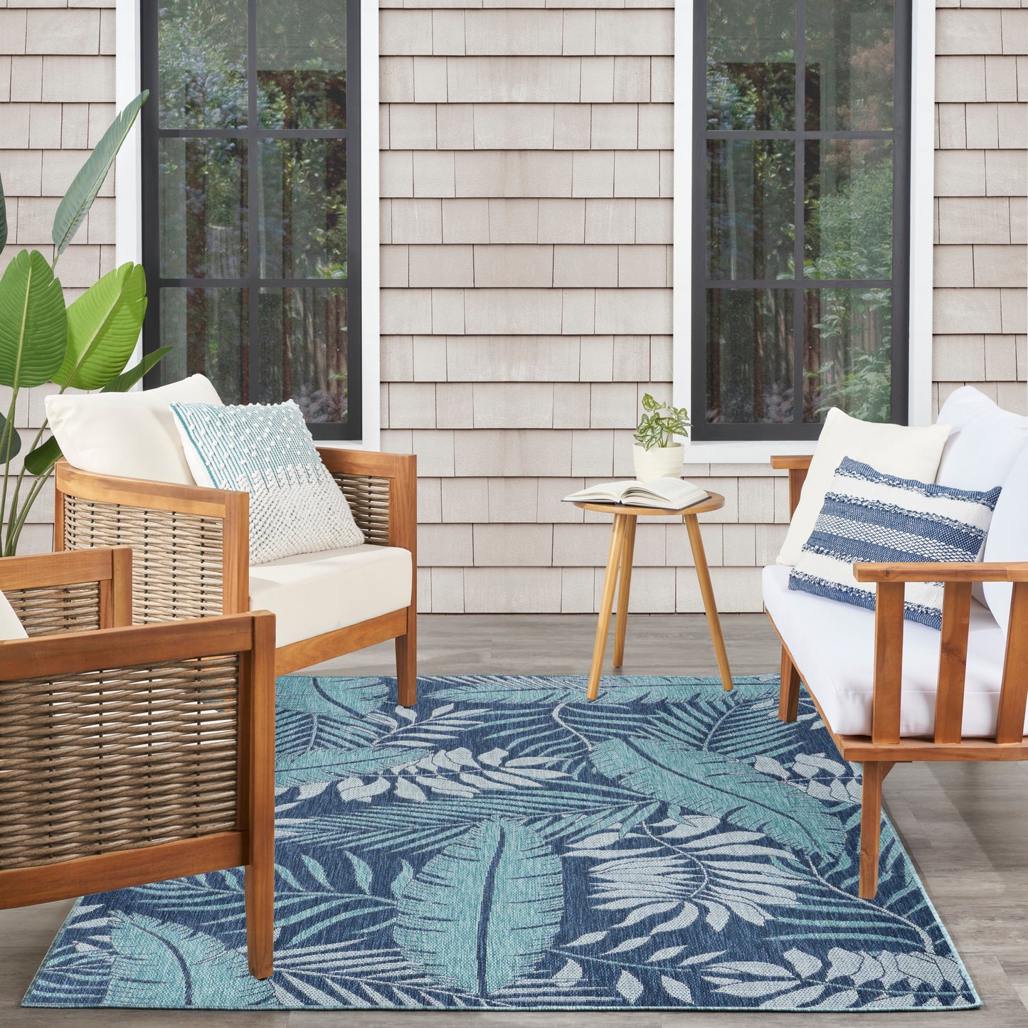 Wood-framed outdoor seating with cream cushions, blue pillows, tropical leaf rug and a small side table with a book