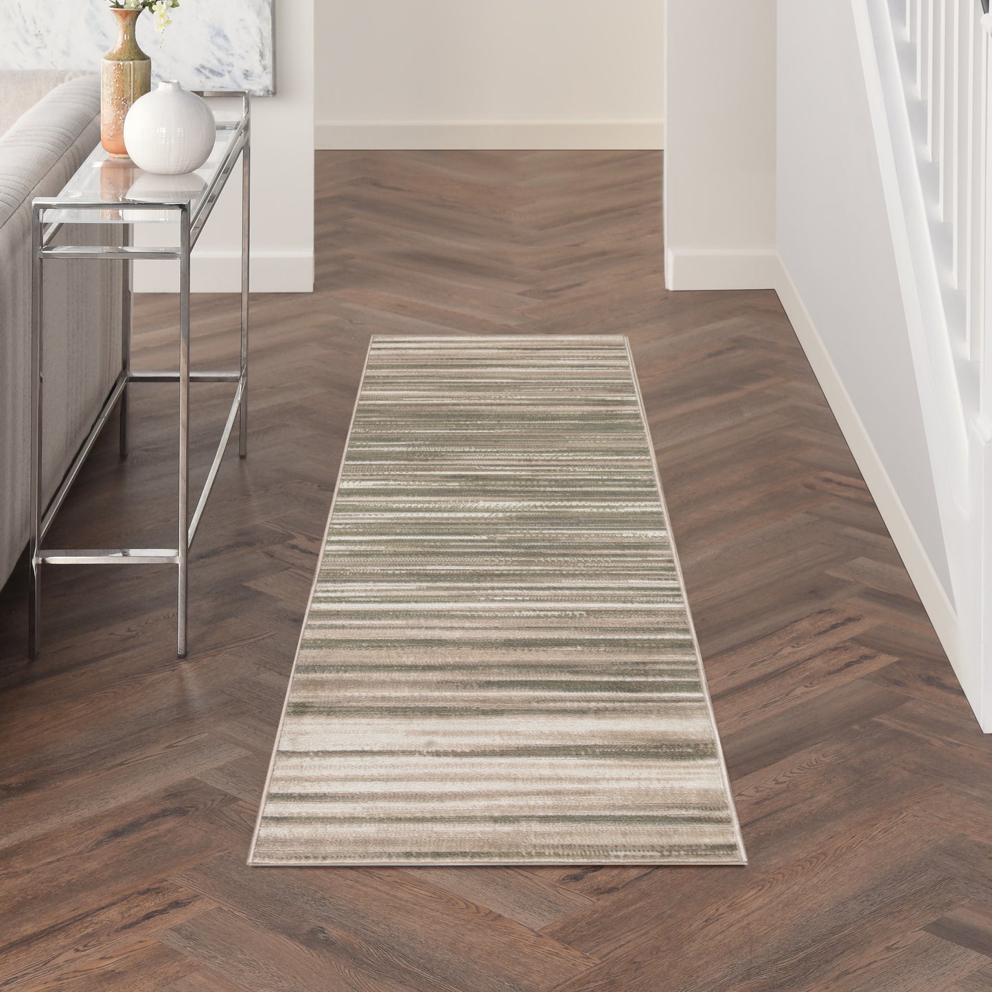 Rectangular beige striped hallway runner rug on dark wood herringbone floor; modern decor with glass console and vase.