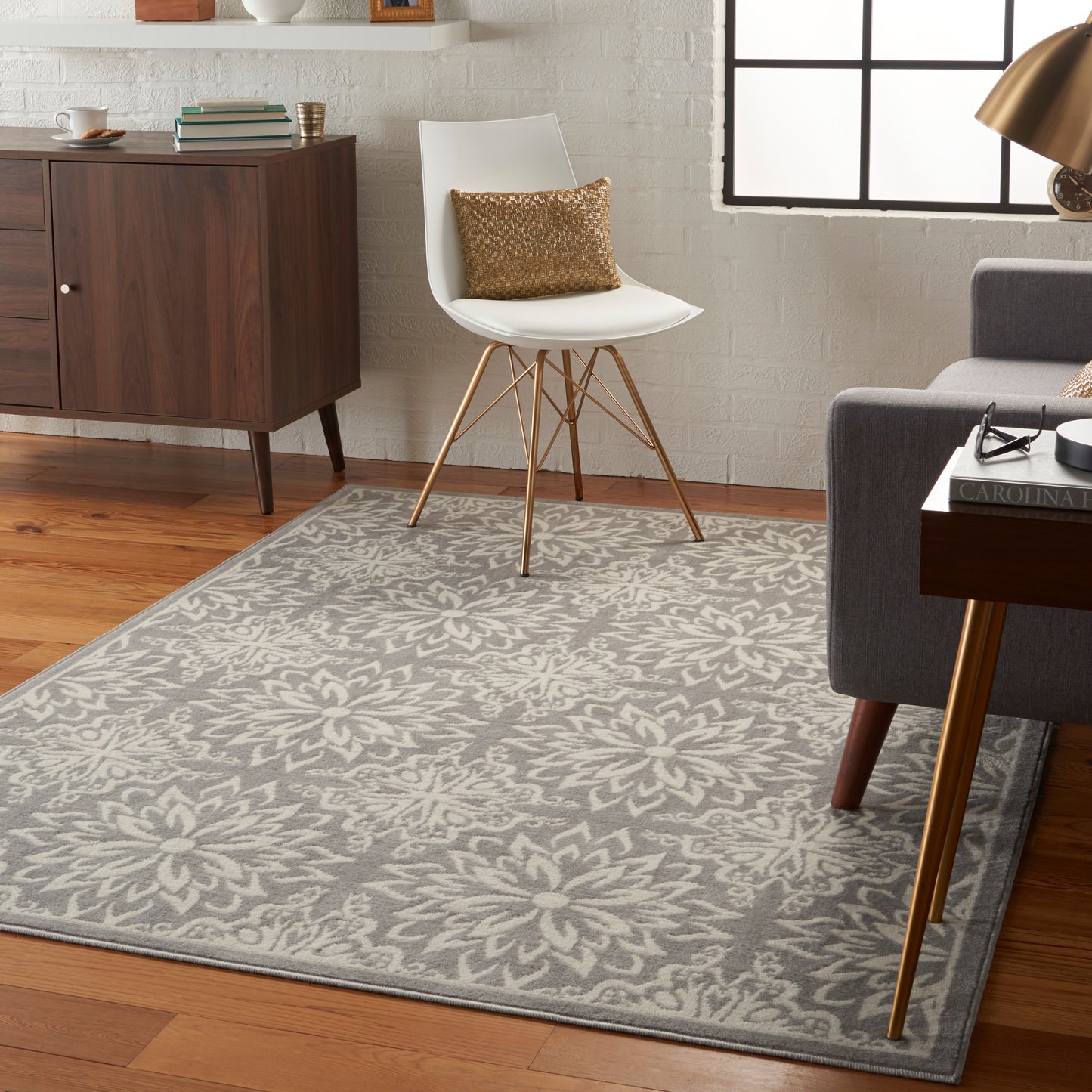 Gray floral-area rug on wood floor with white mid-century chair, gold legs, and a metallic gold throw pillow.