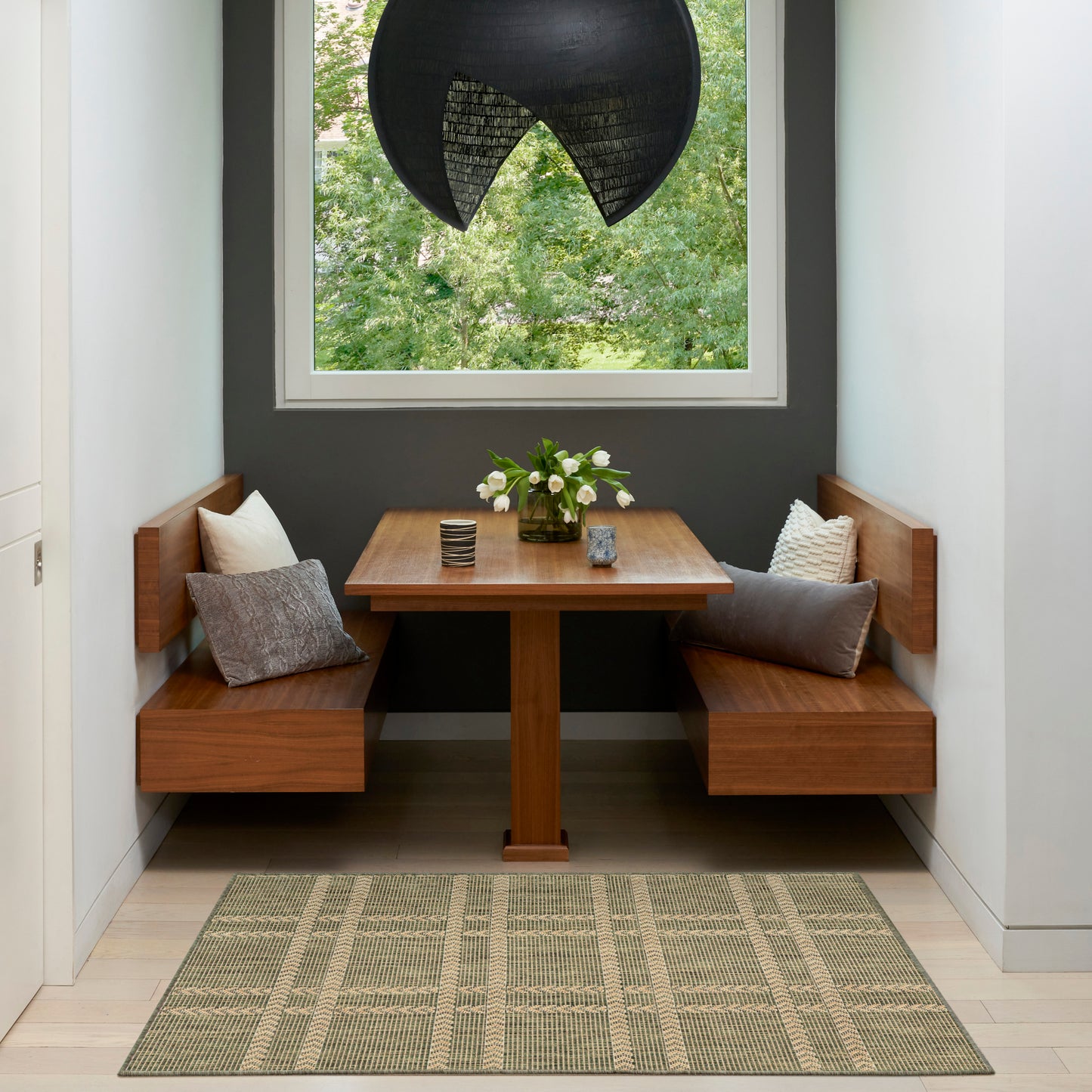 Wooden corner dining set with square table, benches, gray cushions, vase of flowers, black pendant lamp, window view.