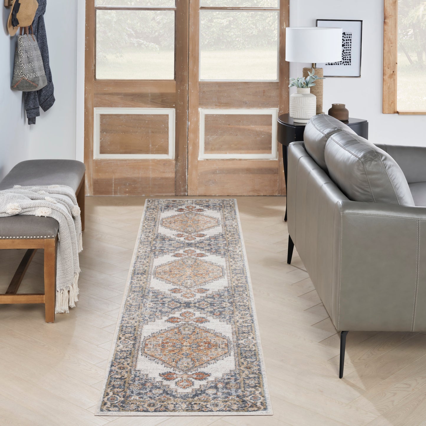 Long runner rug with geometric medallions in ivory, blue, rust on light wood floor beside gray sofa and wooden doors.
