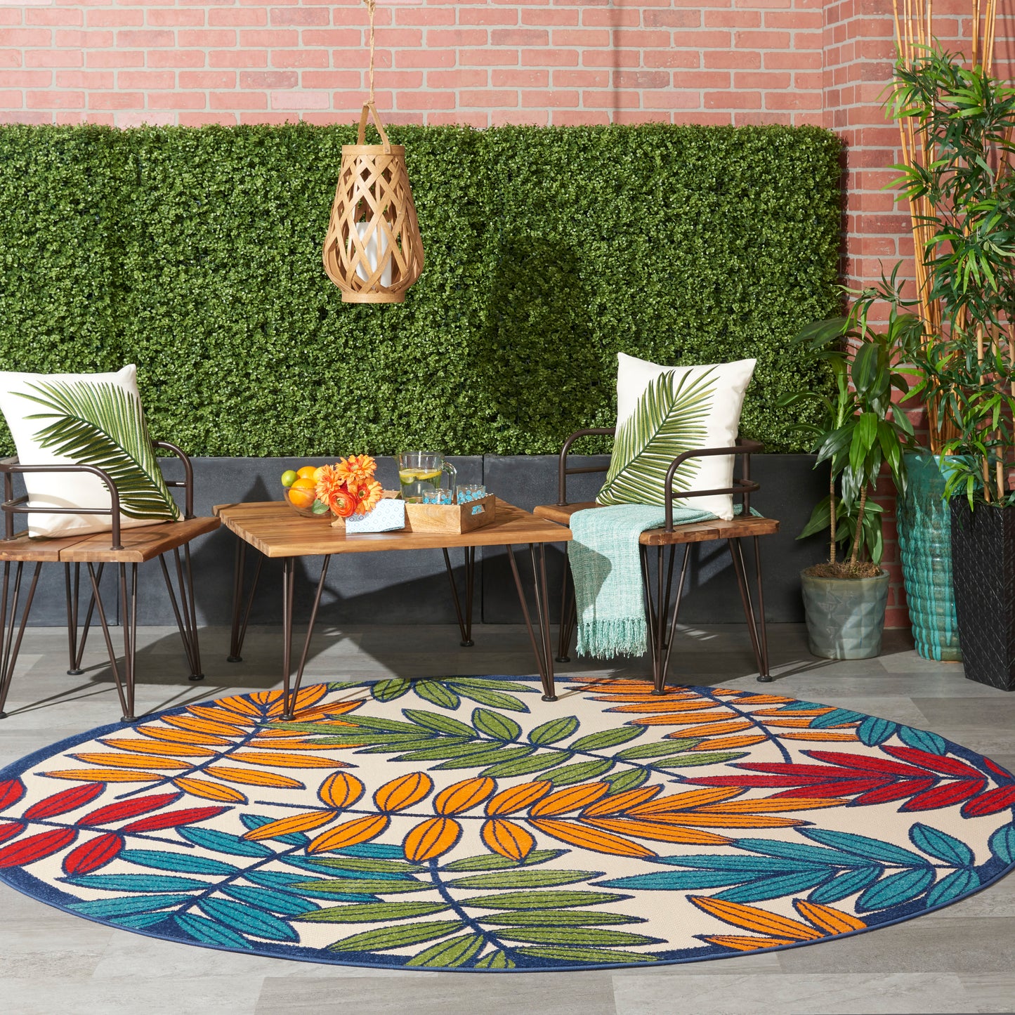 Outdoor patio with round leaf rug, wicker chairs, wooden table, tropical cushions, hanging lantern, plants, brick wall.