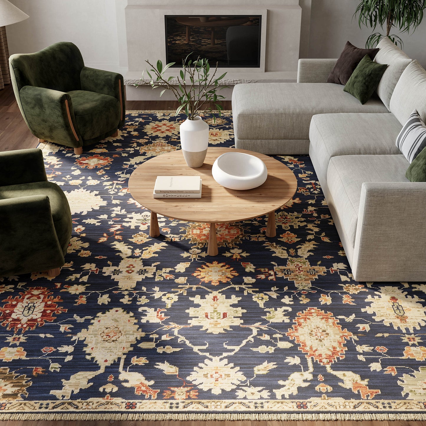 Living room with a navy floral rug, round wooden coffee table, white vase and bowl, beige sectional sofa, and green velvet armchairs.