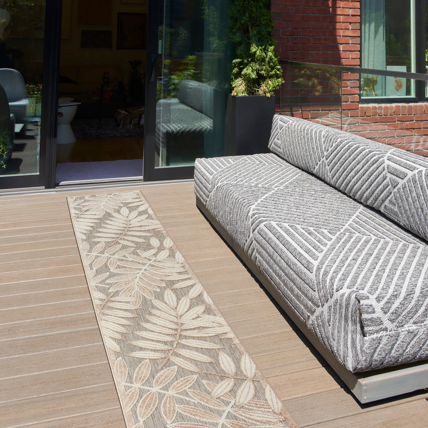 Outdoor lounge: grey patterned modular sofa on a wooden deck with a beige leaf-pattern rug beside sliding glass doors.
