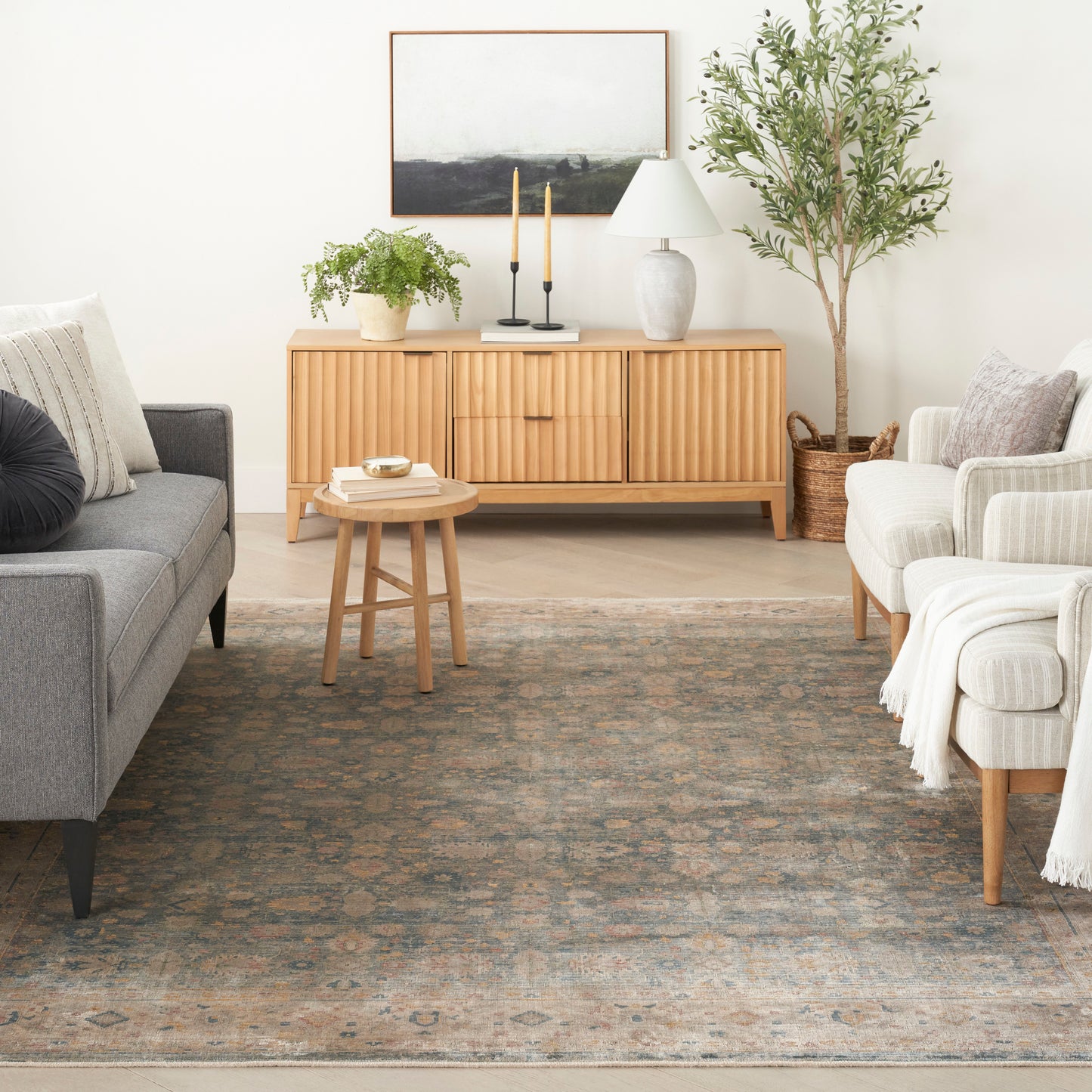 Patterned area rug in a living room with a light wood sideboard, grey sofa, beige armchairs, a potted plant, lamp and candles.