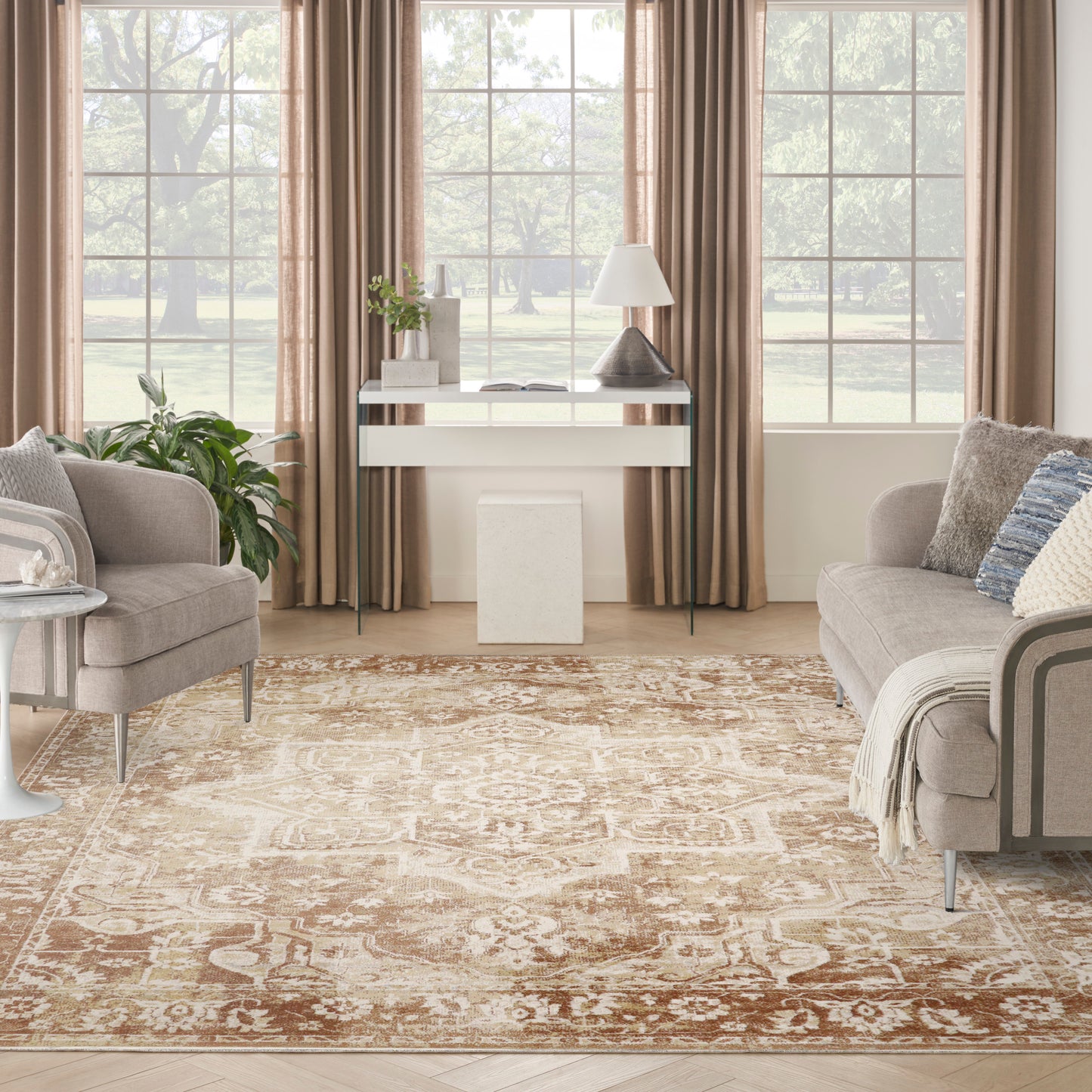 Cozy living room with beige sofas, glass-top console, white lamp, patterned rug, plants, and large windows with tan curtains.