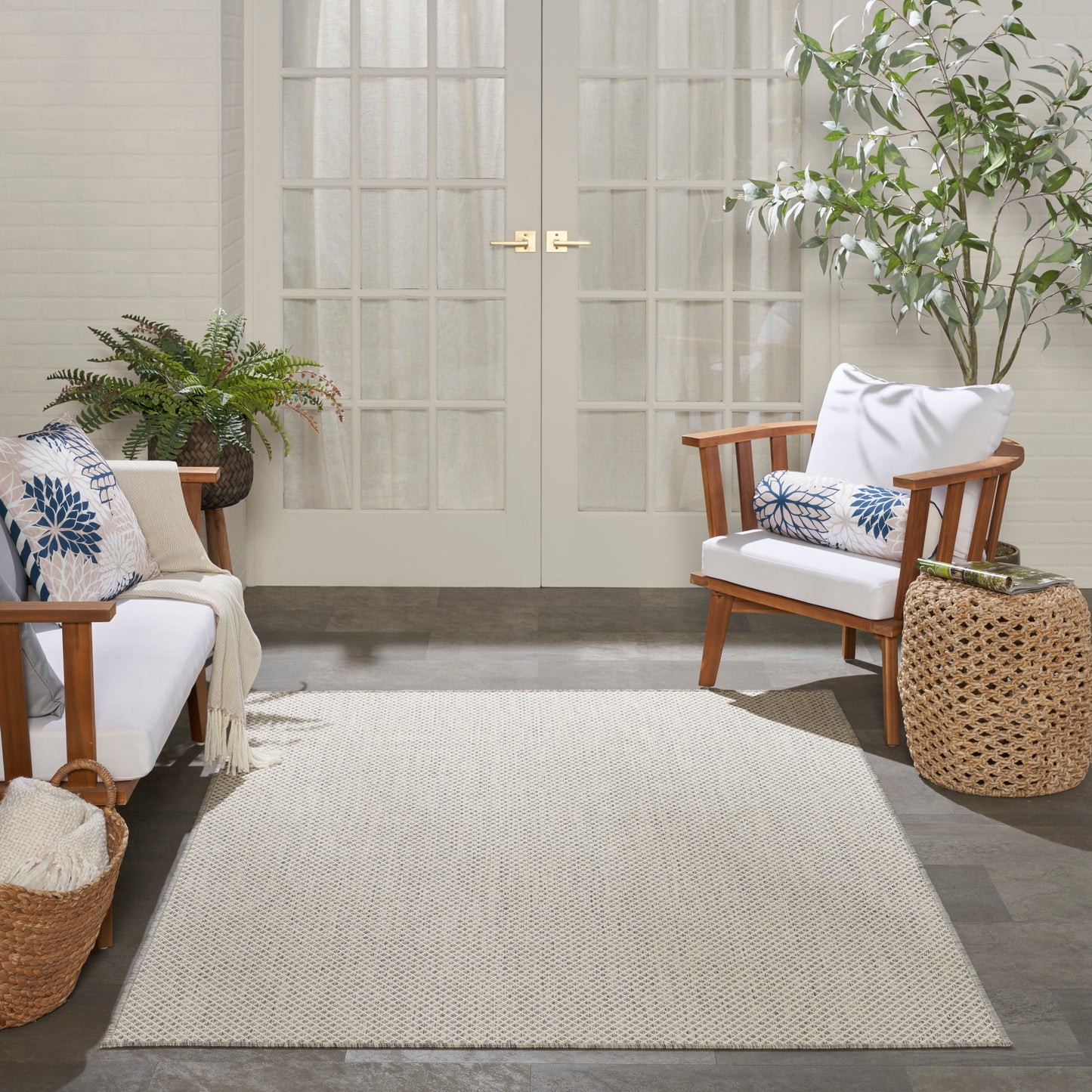 Beige woven rug between two natural wood armchairs with white cushions and blue pillows; plants, wicker basket, French doors.