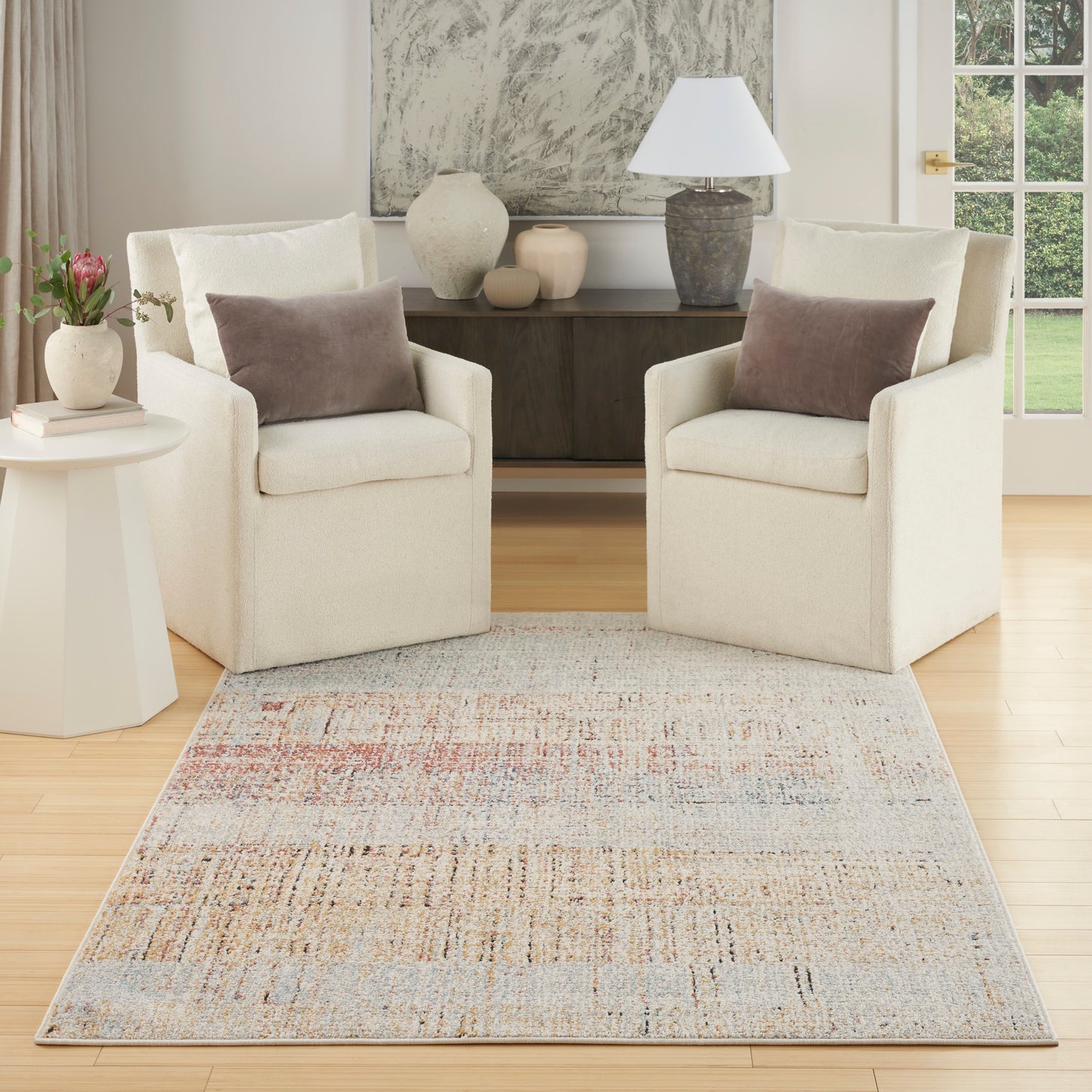 Two cream fabric armchairs with brown pillows flank a dark wood console; pale multicolor rug ties the bright living room.