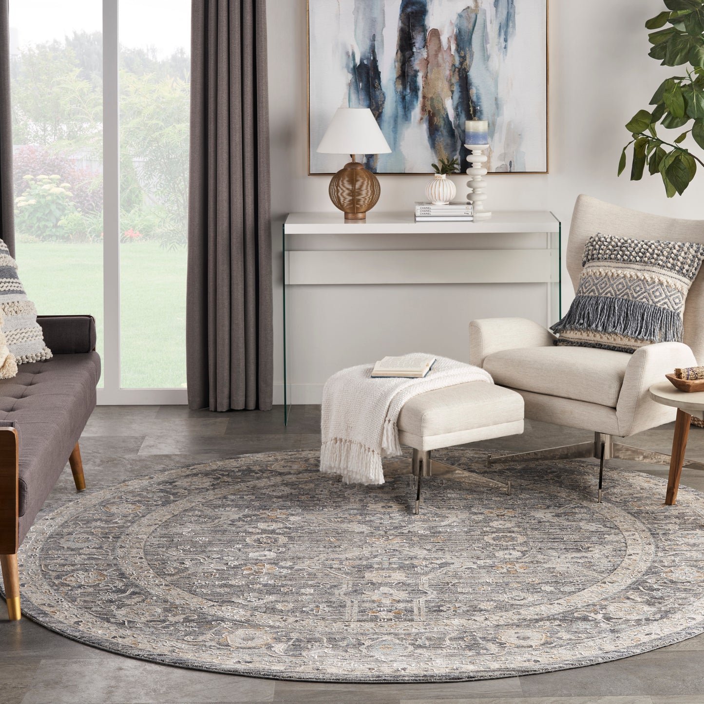 Round gray-blue patterned area rug under beige lounge chair & ottoman with knit throw; glass console & abstract wall art.