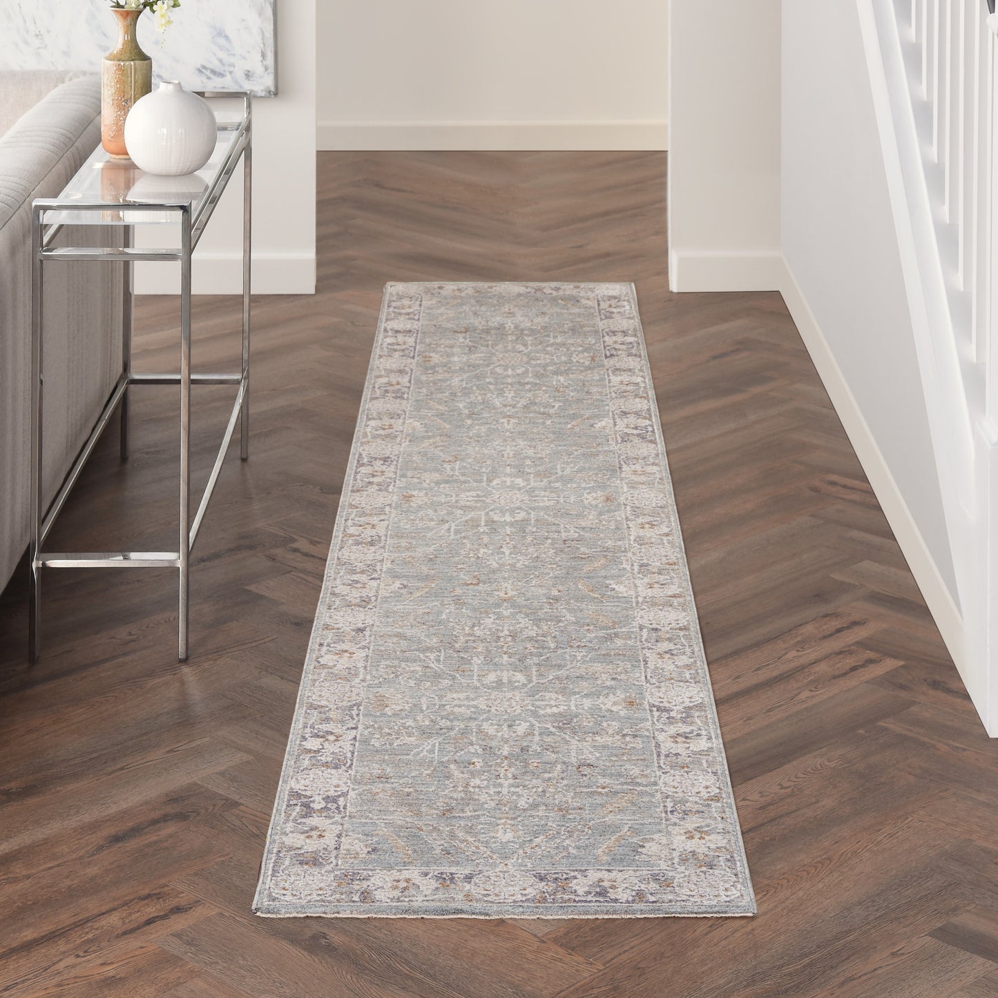 Light gray patterned hallway runner rug with beige accents on dark herringbone floor beside a glass-top chrome console.