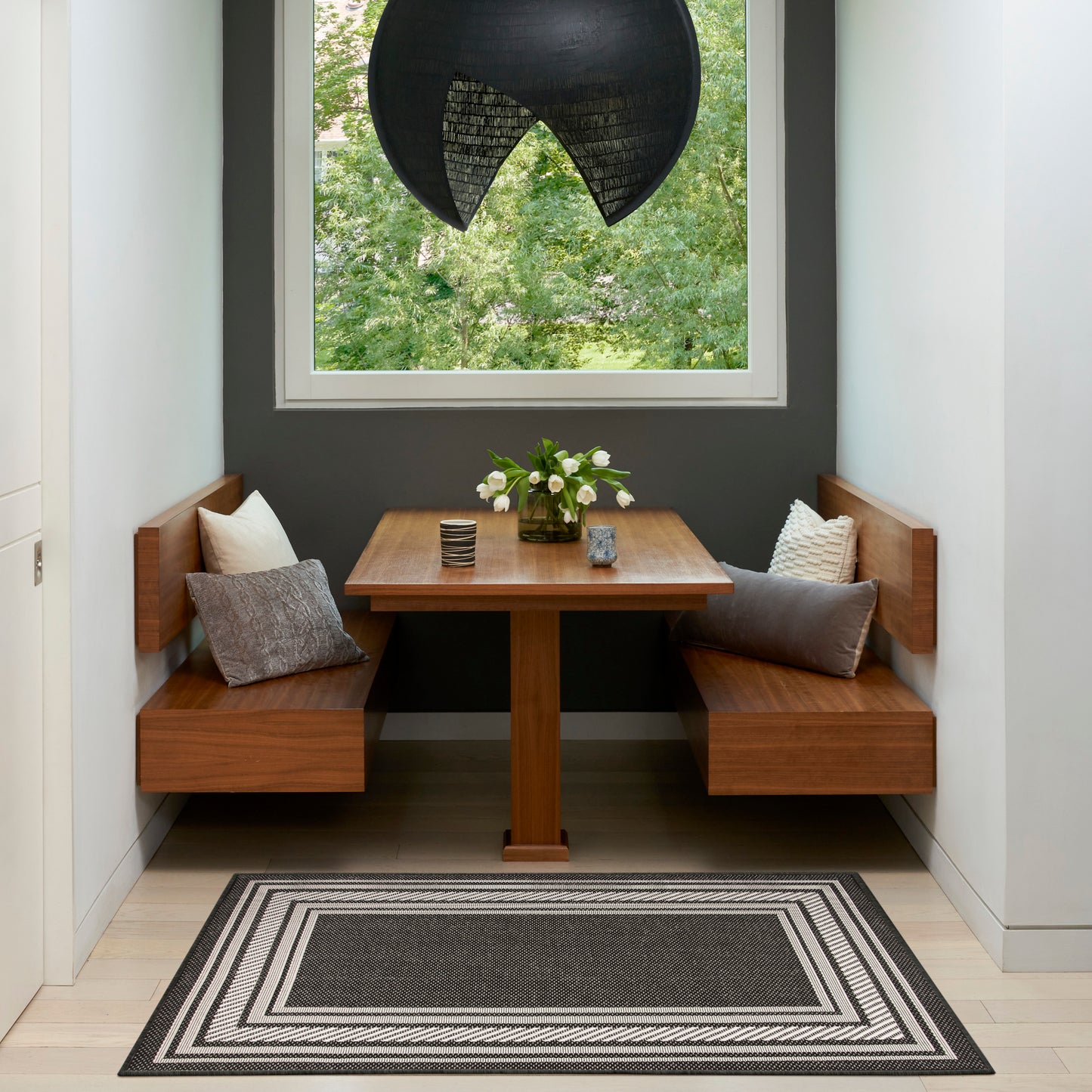 Cozy dining nook: square walnut table with curved benches, gray and white cushions, black pendant lamp, striped rug.